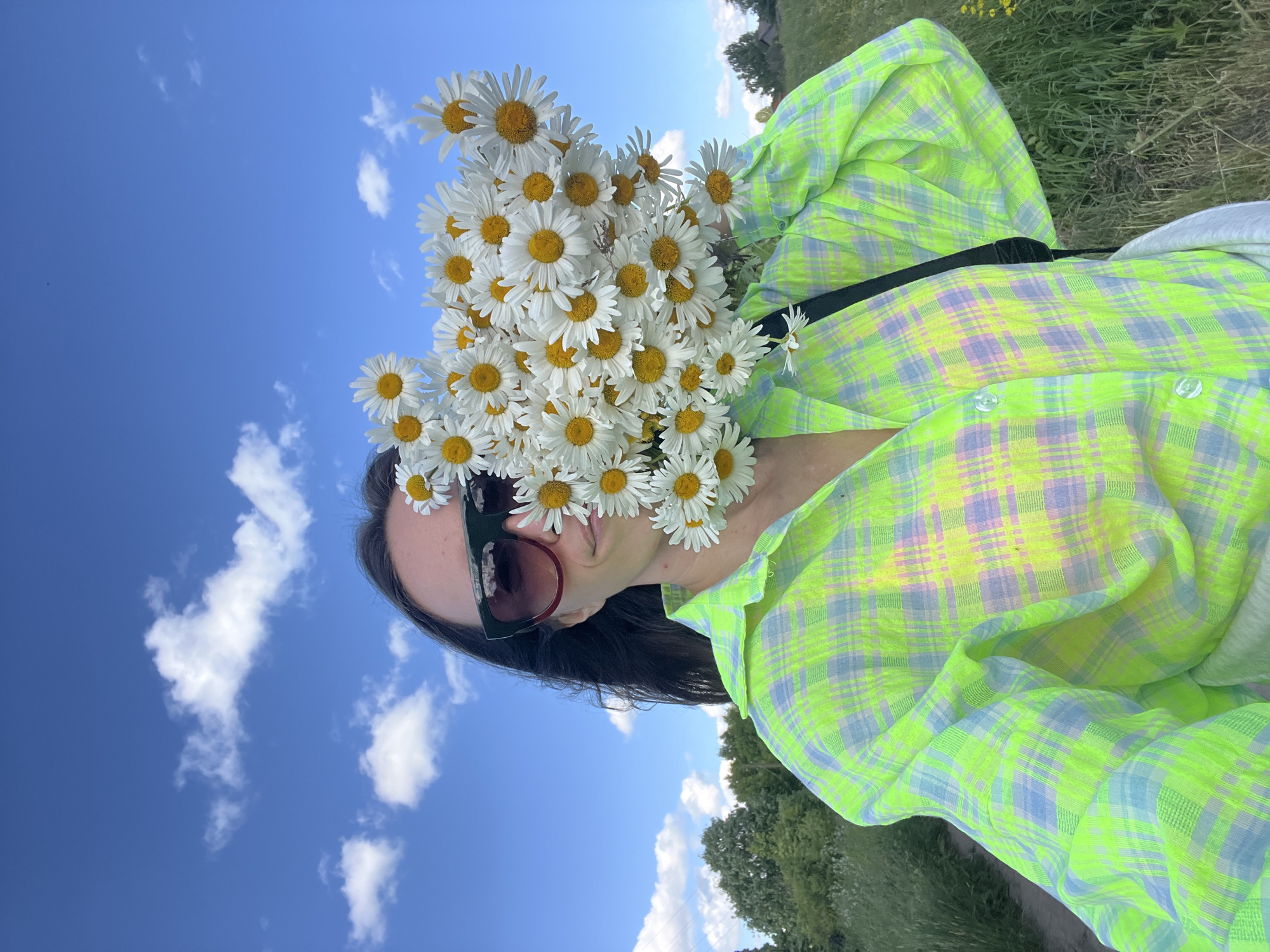 A young woman with dark hair and sunglasses is holding a large bouquet of daisies in front of her face, obscuring most of her features. She is wearing a bright green and purple plaid shirt and is standing outdoors in a field with green grass and trees in the background. The sky is blue with scattered white clouds. The image appears to be a selfie taken on a sunny day. This scene is located in the city of Pigozino, Russia.