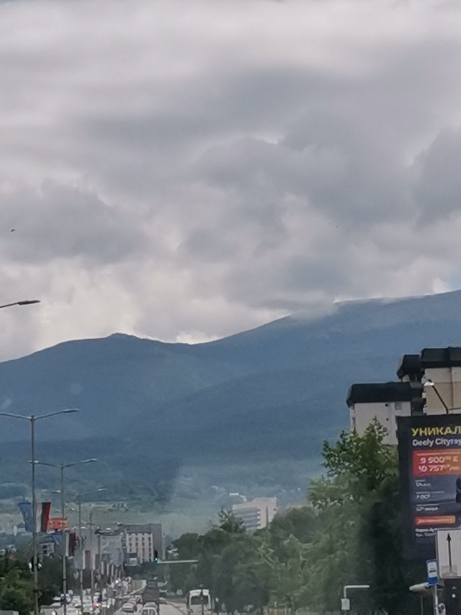 This is a photograph taken in Sofia, Bulgaria, showing a view of Vitosha Mountain under a cloudy sky. In the foreground, there is a street with traffic and several buildings. A billboard advertising "Geely Cityray" is visible on the right side of the image. The mountain appears hazy, with some clouds or mist clinging to its slopes. The overall atmosphere is overcast and somewhat muted.