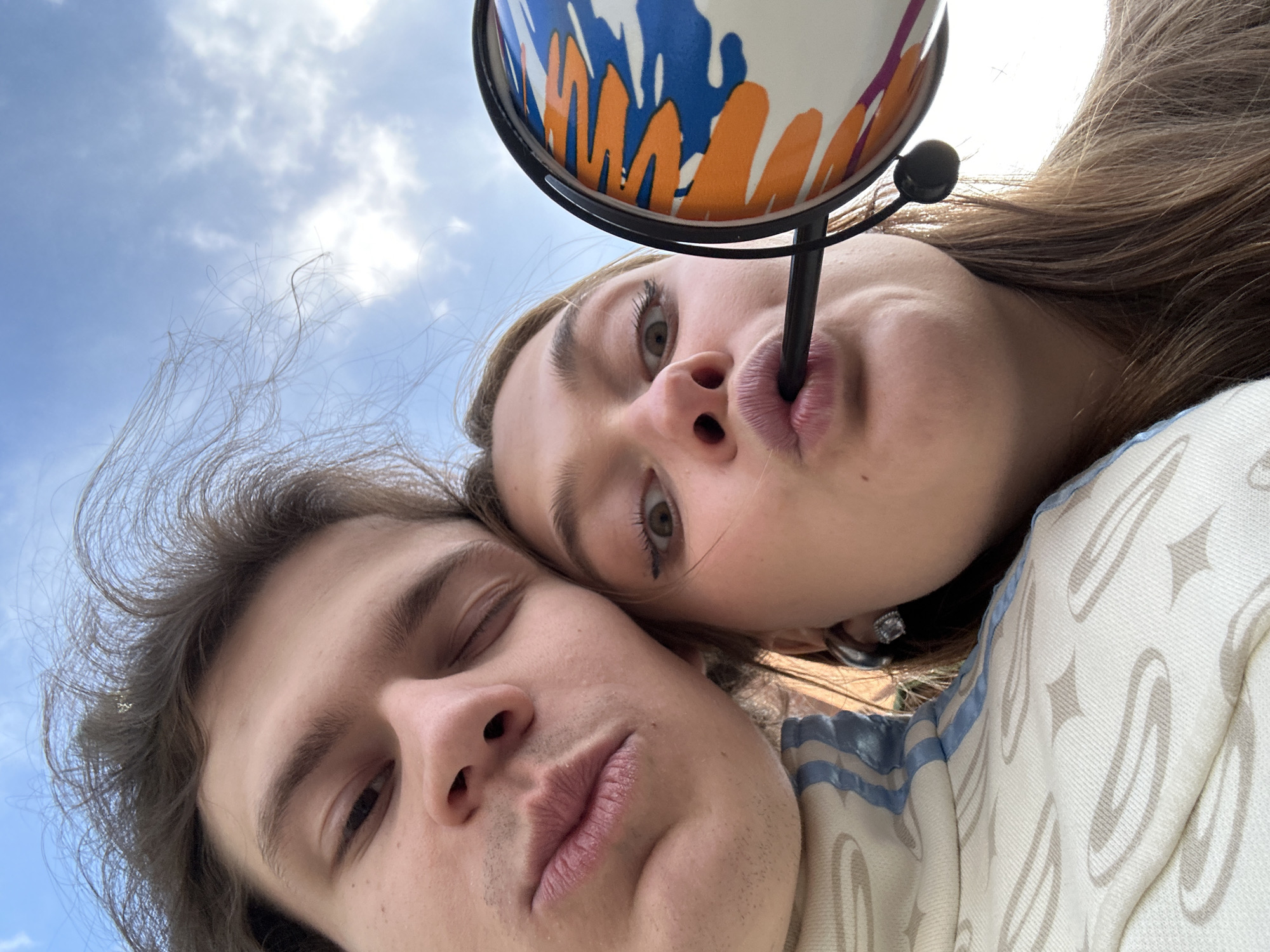 A low-angle selfie captures two people, a man and a woman, in front of a partially visible cloudy sky. The woman, positioned above and slightly to the right of the man, has light brown hair and is drinking from a cup or glass with an orange, blue, and white design using a black straw. She is looking down at the camera. The man, with dark hair, is slightly below and to the left of the woman, with one eye closed and the other open. They are wearing light-colored clothing. The location of the photo in Krasnoyarsk, Russia is unknown.