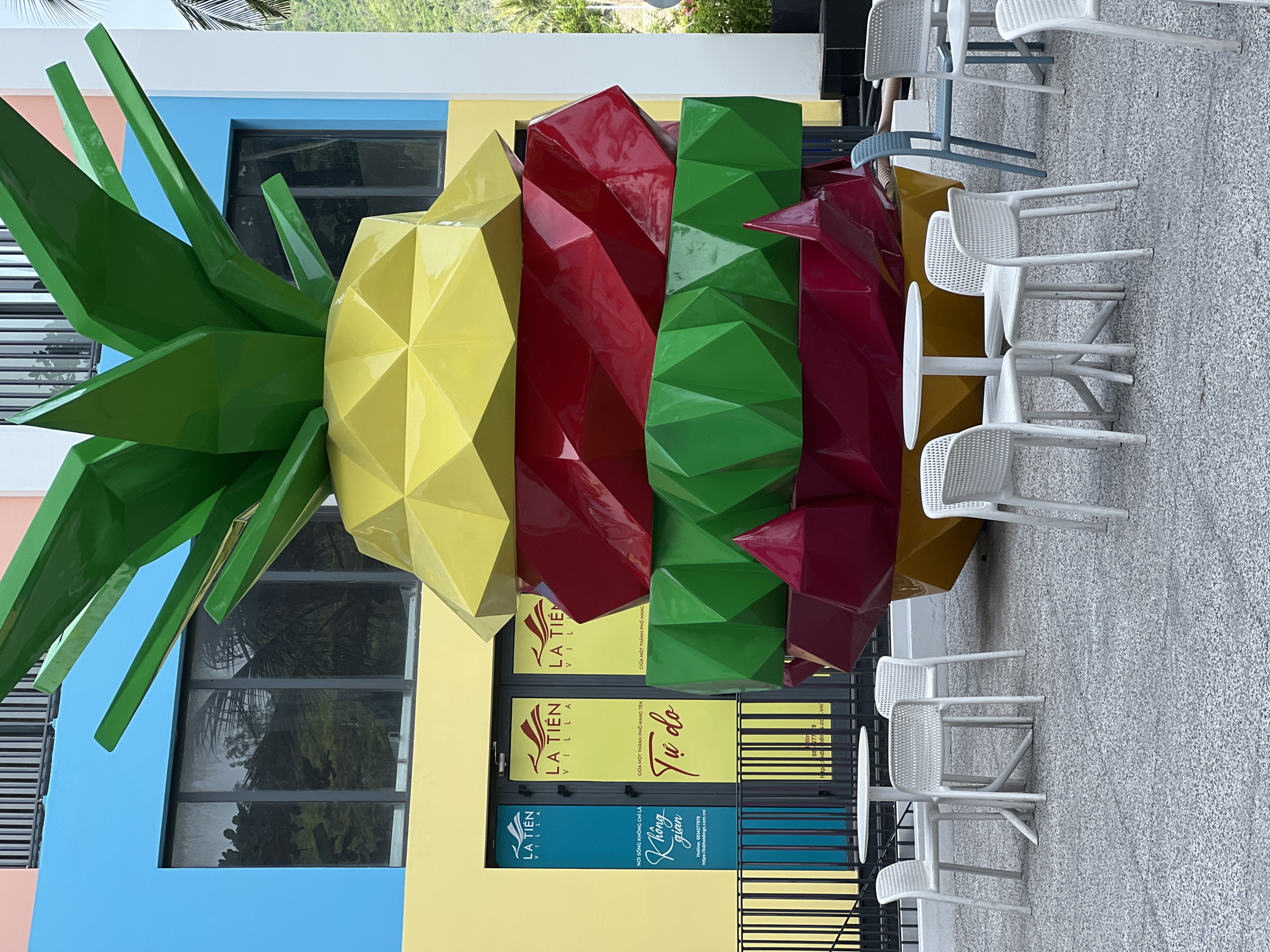 In Nha Trang, Vietnam, an outdoor seating area is captured in front of a vibrantly colored building. The focal point of the scene is a large, geometric sculpture resembling a pineapple, with distinct sections of yellow, red, and green. The building behind it features bright blue and yellow facades, with large windows reflecting the surrounding environment. Several white plastic tables and chairs are arranged on a textured gray ground, suggesting a public space like a cafe or restaurant patio. A black metal fence separates the seating area from the building's entrance, where multiple doors are visible with signage, including "LA TIEN VILLA." In the background, hints of greenery and a hillside can be seen, indicating an urban setting with natural elements. There are no people visible in the frame.