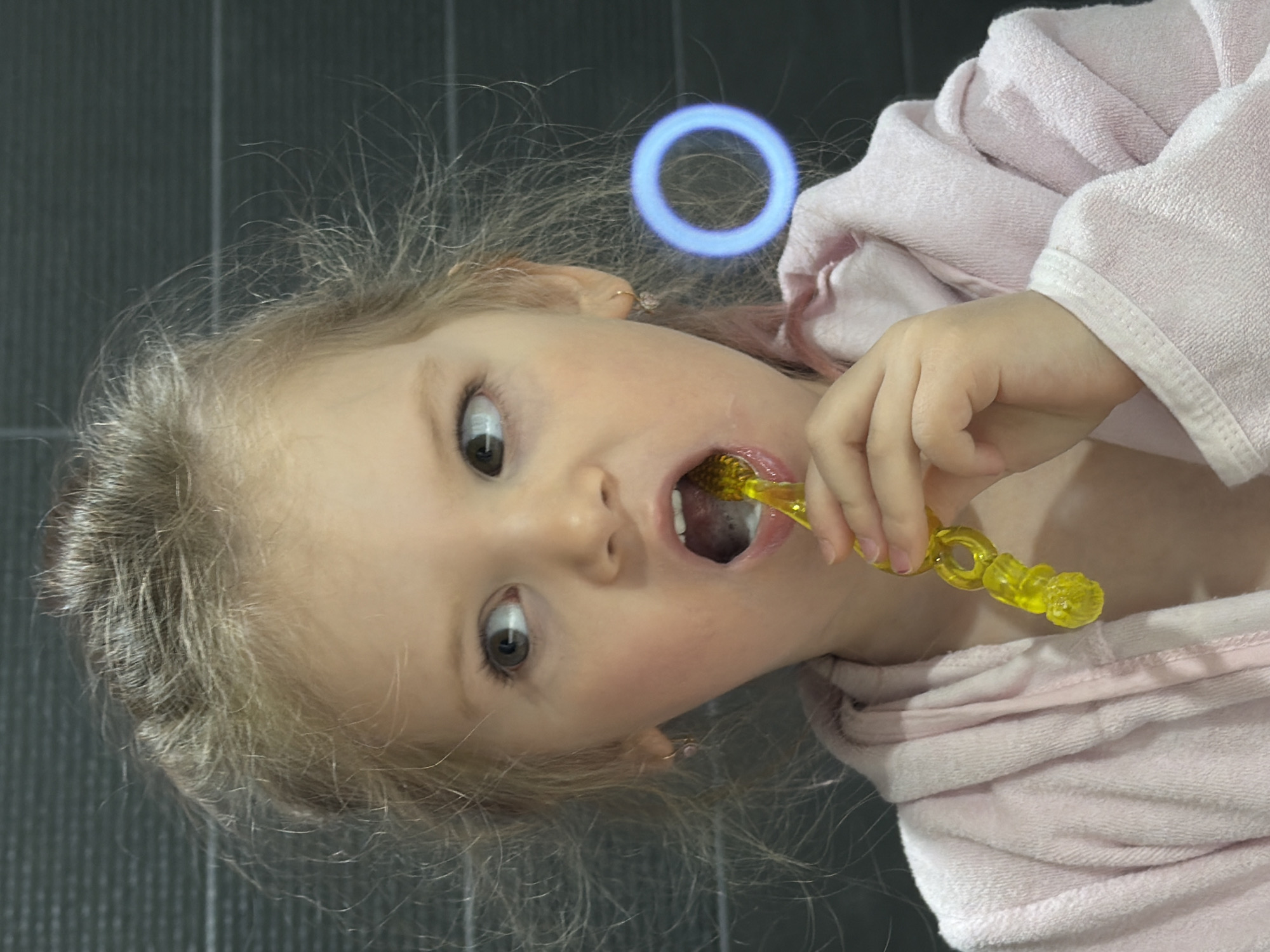 An eye-level shot captures a young child, likely a toddler or preschooler, brushing their teeth. The child has fair skin and light, somewhat messy hair, and is wearing a soft, light pink terrycloth robe. Their eyes are wide and focused, looking directly at the viewer with an expression of concentration or curiosity. A bright, translucent yellow toothbrush, featuring a distinct spiral or textured handle, is held in their hand and positioned inside their open mouth as they brush. The background consists of a dark, vertically-lined surface, possibly tiled, suggesting a modern bathroom environment. A prominent detail in the upper background is a glowing blue circular light fixture, emitting a soft, ethereal light. The image depicts a routine personal hygiene activity and was captured in Chaikovsky, Russia.