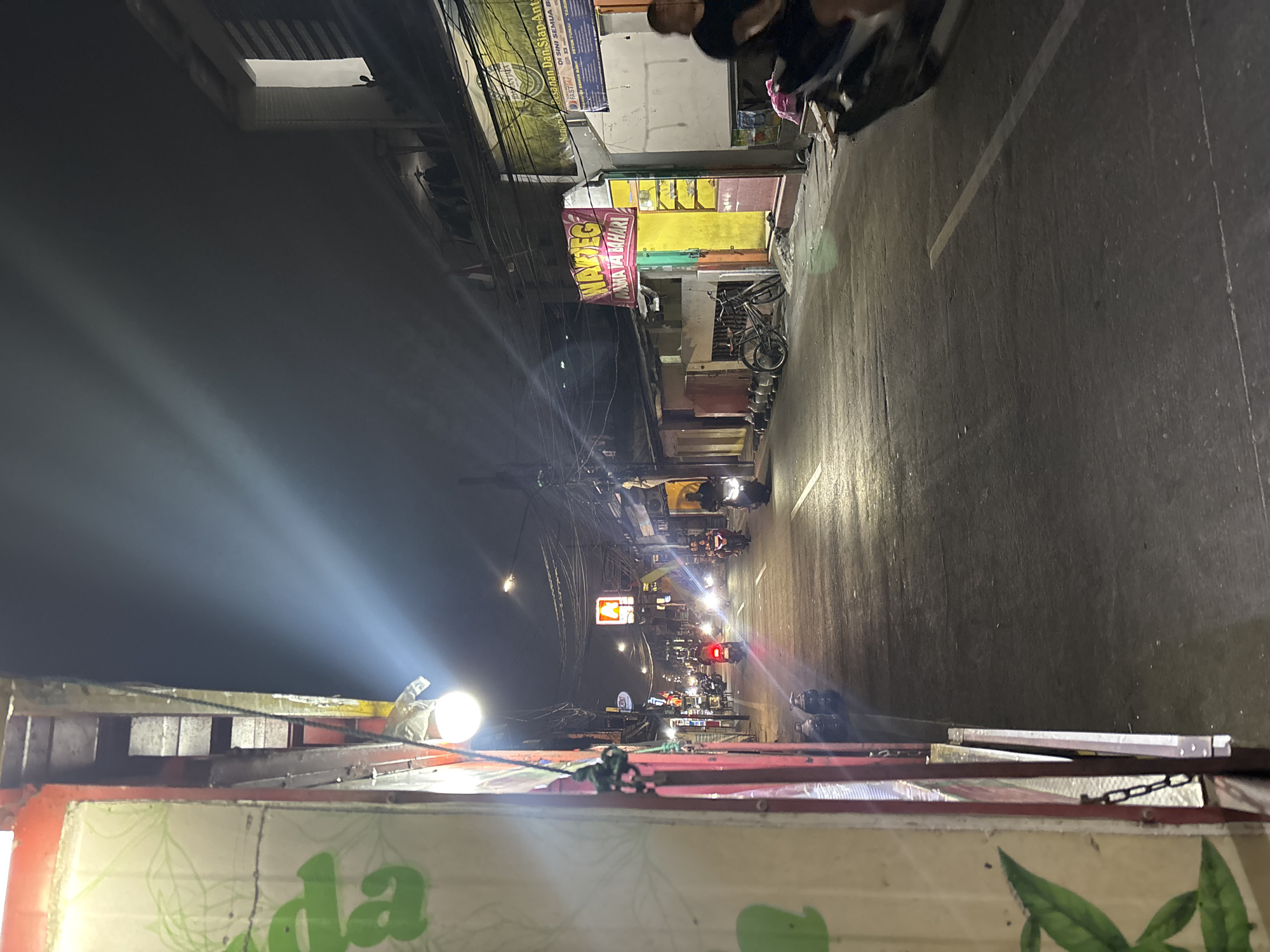 The image captures a nighttime street scene in North Jakarta, Indonesia. A dimly lit street is lined with various establishments, including a "Warung" with a vibrant yellow and green facade displaying the word "WARTEG" in red lettering. A bicycle is parked in front of this establishment. The street is active with several motorbikes on the road, some with illuminated headlights. A person is visible riding a motorbike in the foreground on the right side of the image. Overhead, a network of electrical wires crisscrosses the dark sky, punctuated by the glow of streetlights and neon signs from the businesses. The overall atmosphere is that of a bustling urban environment at night.