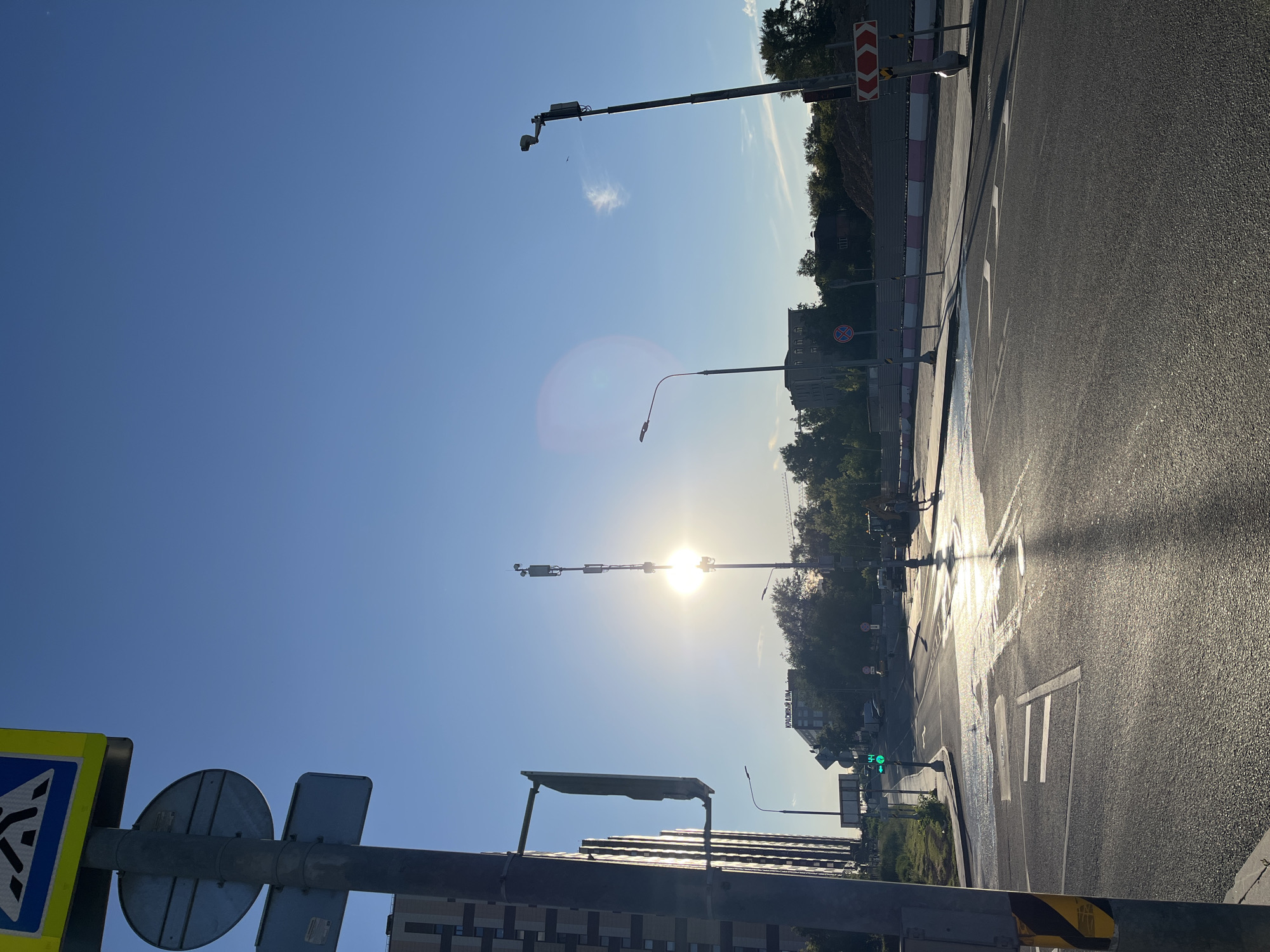 The image depicts a street scene in Moscow, Russia, during a bright, sunny day, likely early morning or late afternoon due to the low angle of the sun. The sun is prominently positioned in the sky, creating a lens flare and casting long shadows across the wet asphalt of a road.
The setting is an urban environment with a clear blue sky. There are multiple traffic lights and street lamps visible, some equipped with cameras. Road markings, including a pedestrian crossing, are clearly visible on the asphalt, which appears to have been recently wet, possibly from rain or street cleaning.
Several buildings line the street, with trees providing greenery along the roadside. A pedestrian crossing sign is visible in the foreground, along with other traffic signs. One sign has red and white chevron markings, indicating a hazard or direction.
The overall impression is one of a typical city street, bathed in the warm glow of the sun. There are no people or vehicles immediately discernible in the image, suggesting a relatively quiet moment. The presence of traffic control equipment and road infrastructure points to a functional urban thoroughfare. One building has text that appears to read "КРАСНЫЙ ДОМ" (Krasnyy Dom), which translates to "Red House".