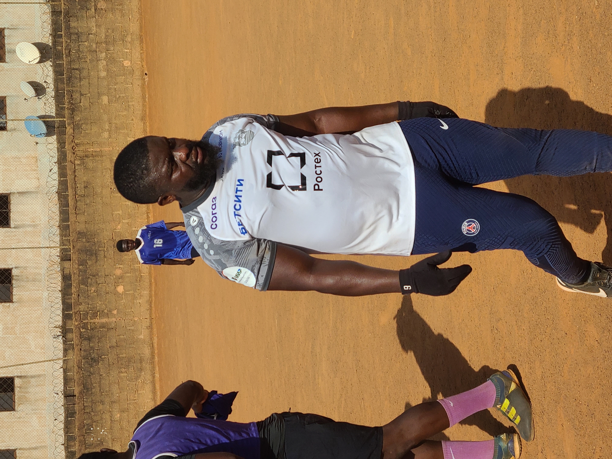 The image depicts a dusty, open area in Barakin Lerawi, Nigeria. A man wearing a white soccer jersey with "Poctex" printed on it, navy blue athletic pants with a Paris Saint-Germain logo, and black gloves is centrally positioned. He is standing on a dirt surface.
In the background, there is a brick wall topped with barbed wire, next to a building with satellite dishes attached. Another man wearing a blue soccer jersey with the number 16 is visible in the background near the wall.
In the foreground, at the bottom of the image, the legs of another individual wearing pink socks and athletic shoes are visible.