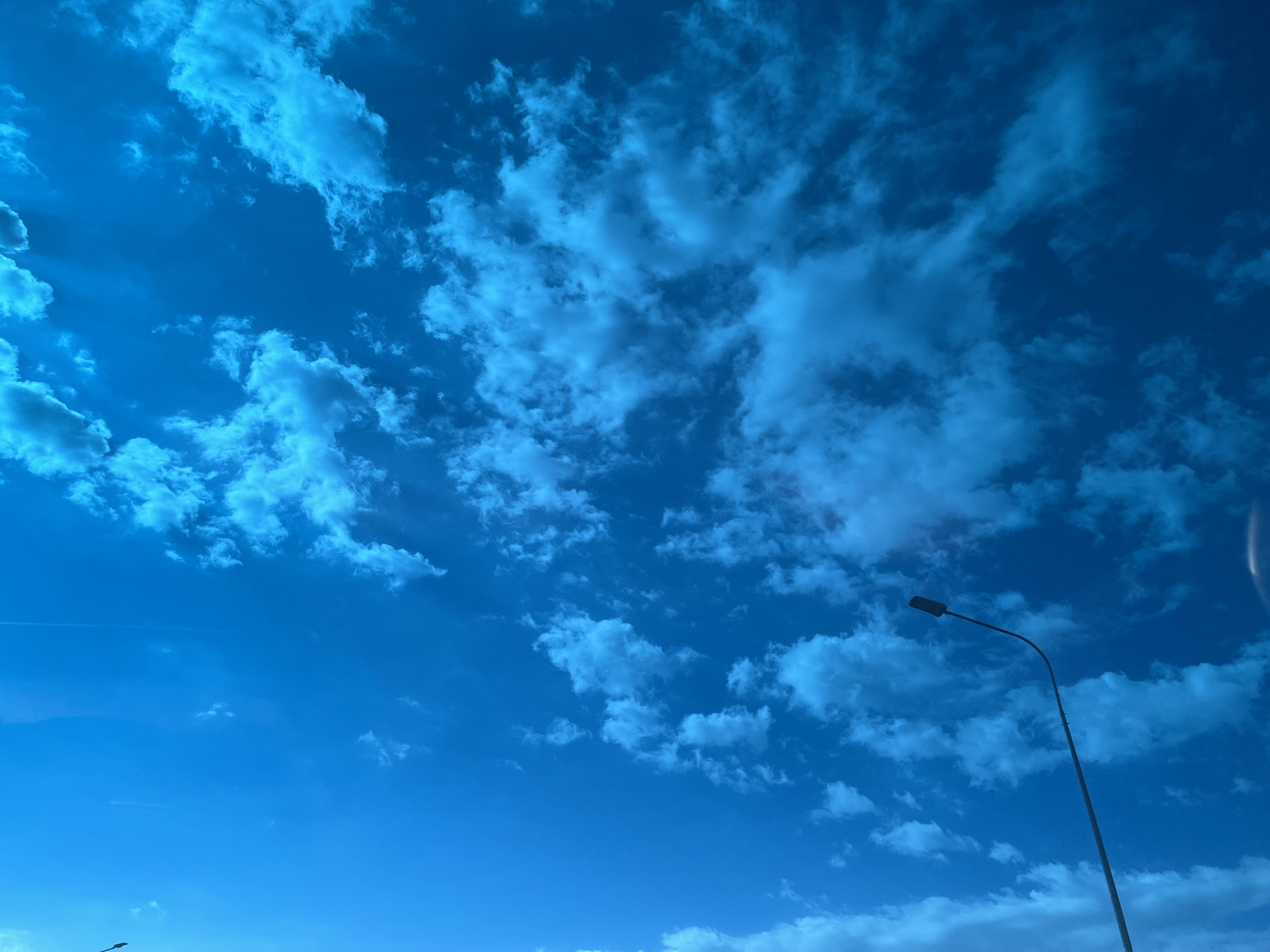 The image depicts a daytime sky, predominantly blue, populated with scattered, puffy white clouds. A tall street lamp extends from the bottom right corner towards the top right. A vapor trail is present in the upper left quadrant. The location is Tugurgoy, Russia.