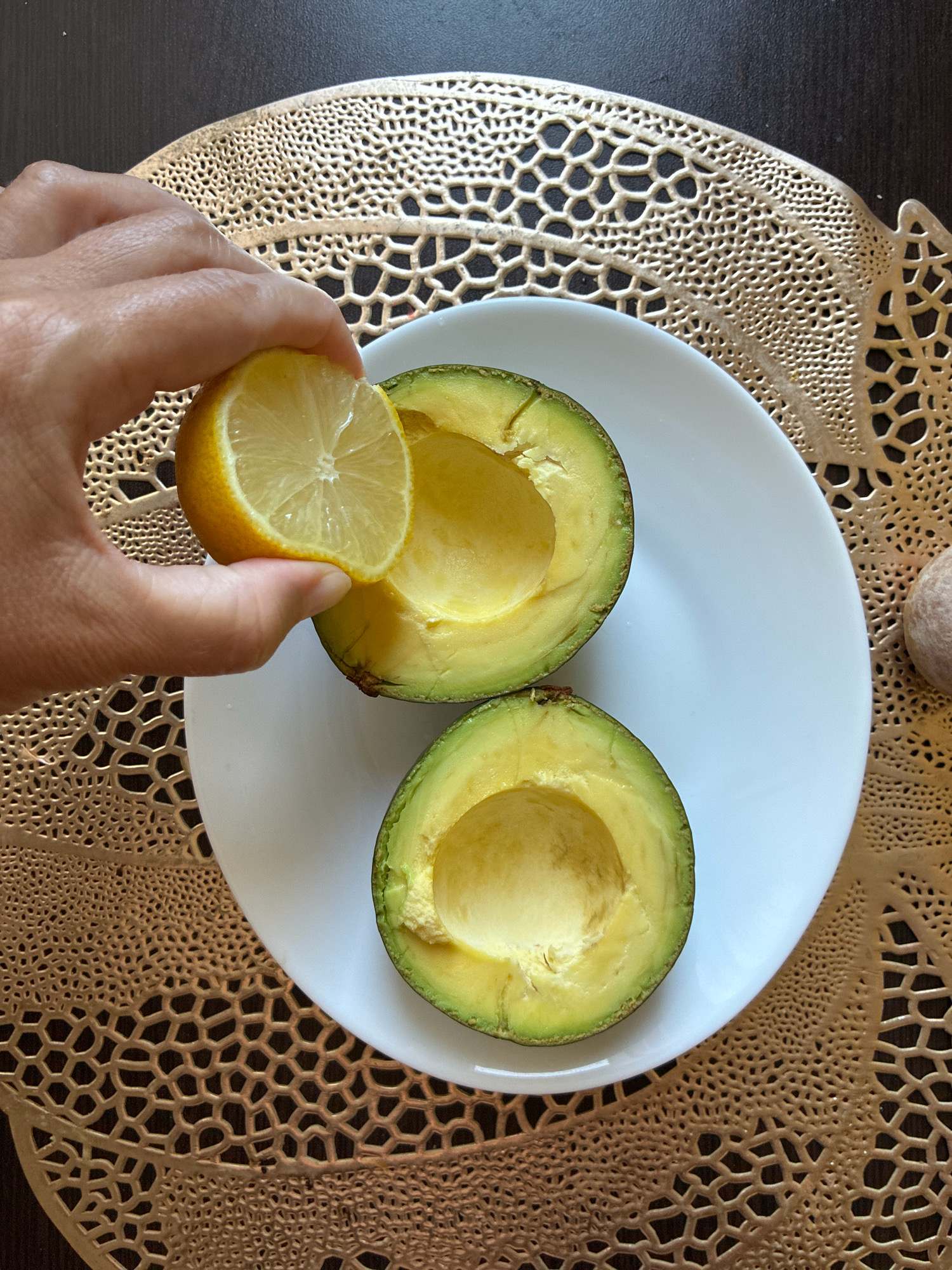 A hand holds a slice of lemon over a halved avocado on a white plate. The avocado is cut lengthwise and placed cut-side up on the plate. The plate is on a gold, ornate placemat, which is on a dark brown surface. The scene is lit from above, creating shadows. No specific location in Sochi, Russia is depicted.