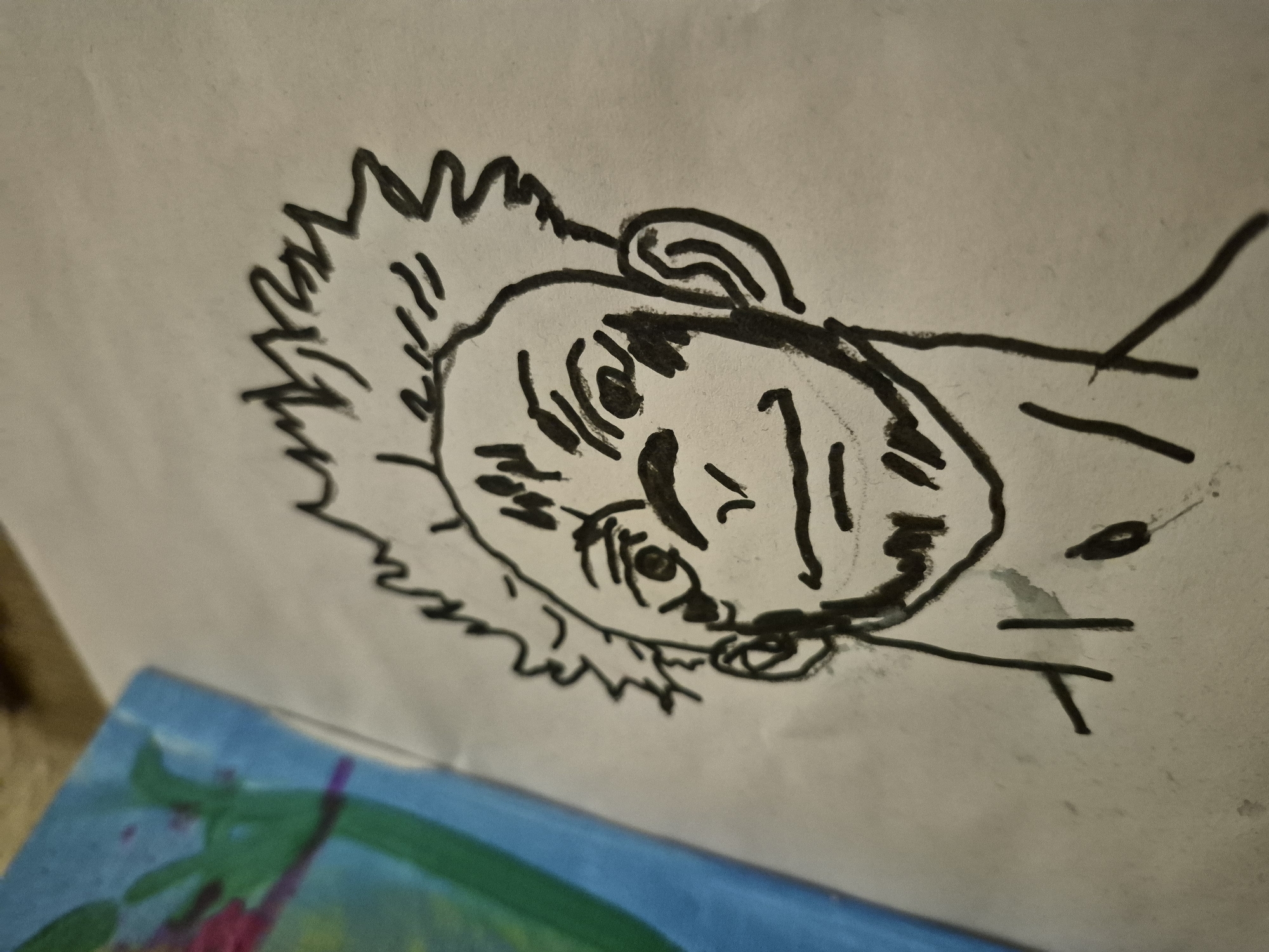 A black and white sketch of a person's head and shoulders is centered on a piece of white paper. The sketch depicts a person with spiky hair, furrowed eyebrows, a large nose, and a goatee. They are wearing a dark shirt. To the left of the sketch, a portion of a colorful drawing is visible, featuring blue, green, and pink hues. The paper is slightly crumpled and textured.
