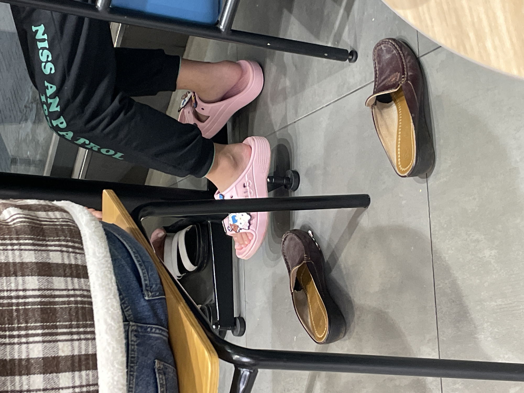 The image, oriented sideways, captures a low-angle view of an indoor setting, likely a cafe or lounge, focusing on the feet and lower legs of individuals seated under tables. One person wears black sweatpants with bright green text reading "NISS AN PA TROL" vertically down the side, suggesting "PAW PATROL" or a similar phrase. This individual's feet are in pink platform slide sandals, one adorned with a white cartoon character charm. Nearby, a pair of dark brown, moccasin-style shoes rests on the light grey tiled floor, indicating the presence of another person. Partially visible in the bottom left corner, a brown and white plaid jacket with a white fluffy lining is draped over a chair, alongside a denim jacket and a wooden furniture piece, hinting at a third person or their belongings. Black metal chair frames and legs are visible throughout the image, defining the furniture. The floor is composed of light grey tiles. The scene depicts people relaxing or waiting in a casual indoor environment, consistent with a setting in Phường Xuân Hương - Đà Lạt, Vietnam.