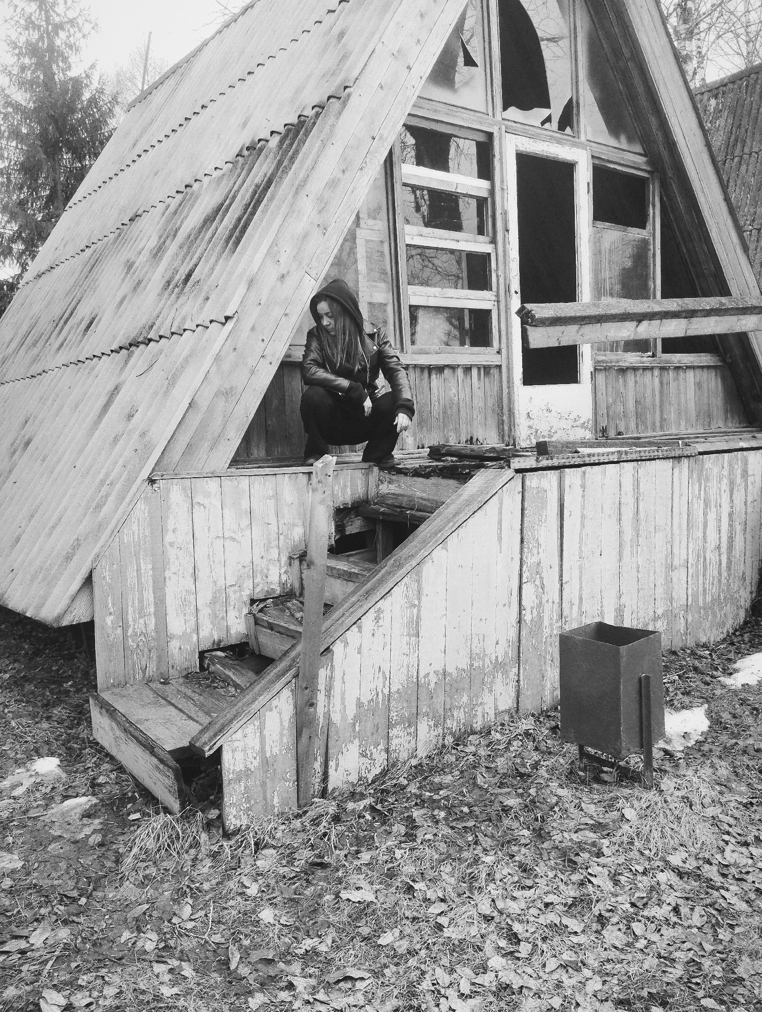 The image, in grayscale, shows a person crouching on the porch of a dilapidated A-frame structure. The structure appears abandoned, with broken windows and peeling paint. The person is wearing a hooded jacket and dark clothing. The stairs leading up to the porch are weathered and partially broken. A metal trash receptacle stands to the side of the stairs. The ground is covered with dead leaves and some patches of snow. Based on the association of the image with the location "Galevo, Russia," it is likely that the structure is located in or near that city.