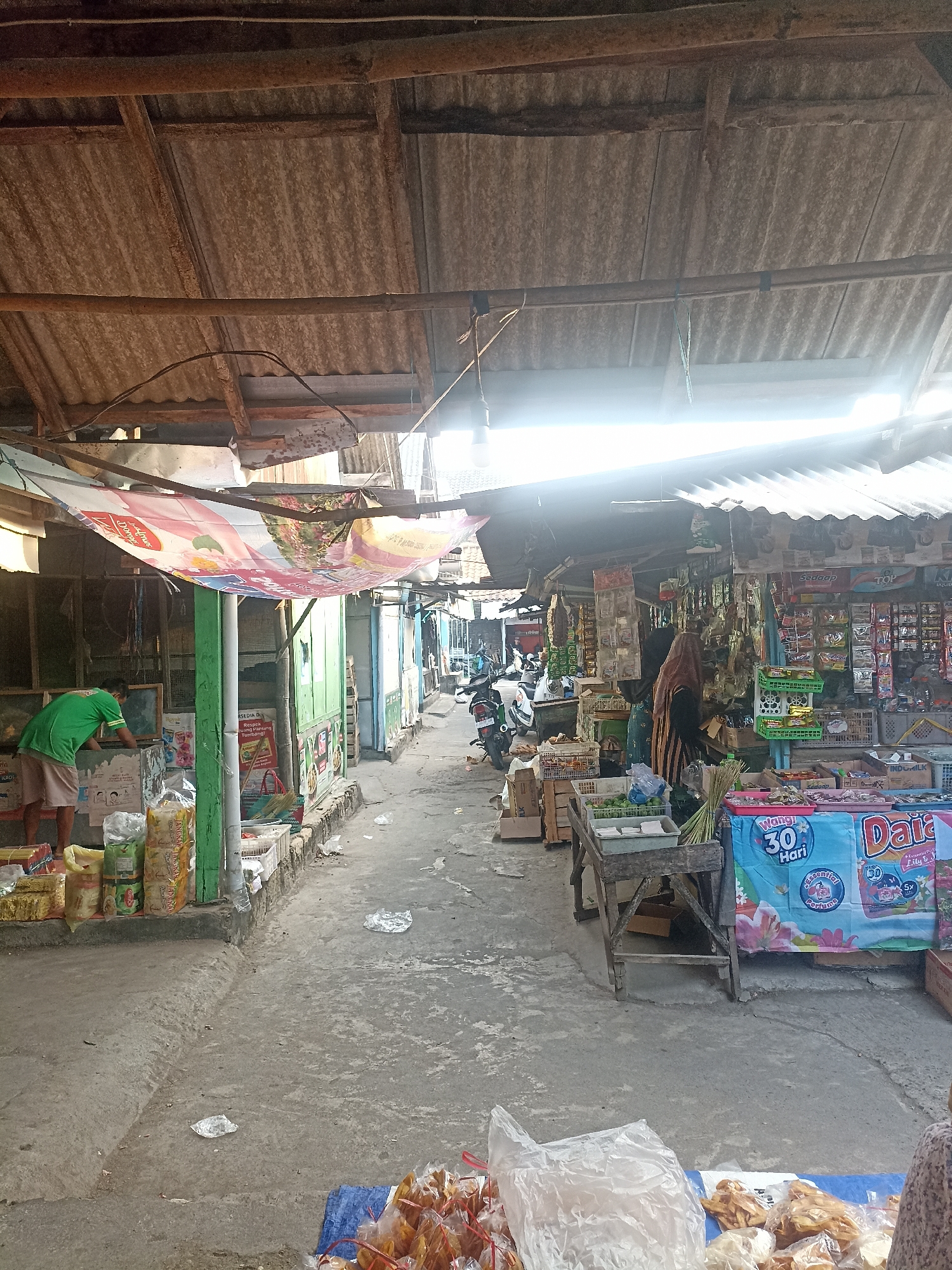The image depicts a market scene in Mantup, Indonesia. A narrow alleyway is lined with stalls on both sides, covered by corrugated tin roofs supported by wooden beams and poles. The ground is paved with concrete.
On the left side of the alley, a person wearing a green t-shirt and light-colored shorts is seen bending over a stall, seemingly organizing goods. In front of the stalls, various packaged food items are displayed, including bags of snacks and what appear to be noodles. A large banner, partially obscured, is hung overhead, displaying colorful graphics and text.
On the right side of the alley, more stalls are visible, packed with a variety of products, including packaged food items like snacks, beverages, and household goods. Two individuals are present near one of the stalls; one is wearing a hijab and a long dress, while another person, partially visible, is also wearing traditional clothing. Several motorcycles are parked along the alleyway, suggesting modes of transportation for vendors or customers. In the foreground of the right side, a display table holds packaged snacks and other items for sale, with a colorful banner featuring floral imagery and text, including "Wangi 30 Hari."
The overall environment appears to be an open-air market, characterized by its rustic construction, the abundance of merchandise, and the presence of people engaged in commercial activities. The lighting suggests it is daytime, with sunlight filtering through gaps in the roofing, creating some bright areas and shadows. The ground shows some debris and litter.