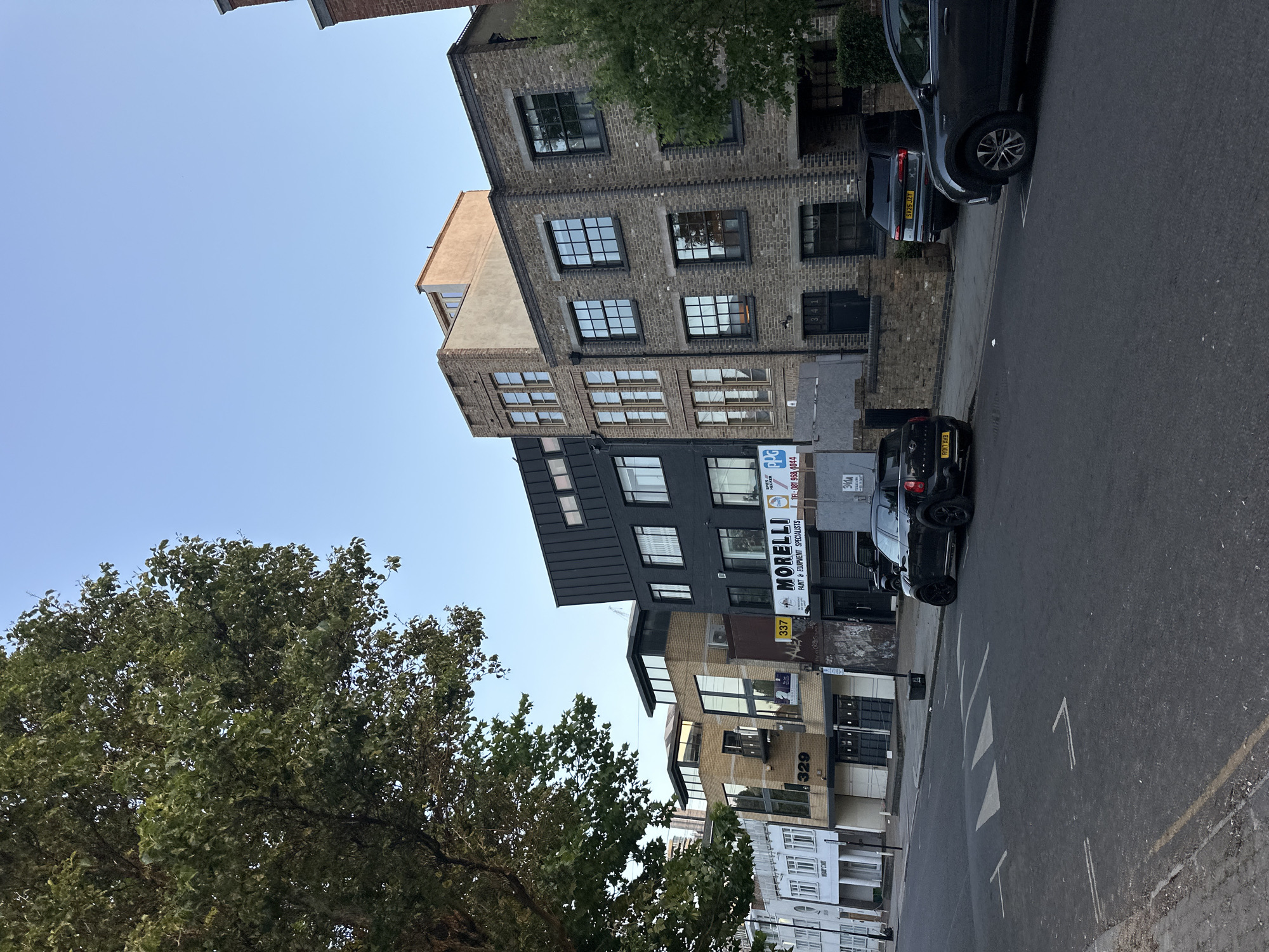 The image shows a street scene in London, United Kingdom. A variety of architectural styles are visible in the buildings lining the street, ranging from brick warehouses to modern residential apartments. A large tree with dense foliage is in the left of the frame. Parked cars line the side of the road. The businesses visible include "MORELLI PAINT & EQUIPMENT SPECIALISTS," indicated by a sign on one of the buildings, and there are numbered units such as 329, 337, and 341. Pedestrian crosswalk markings are visible on the street.
