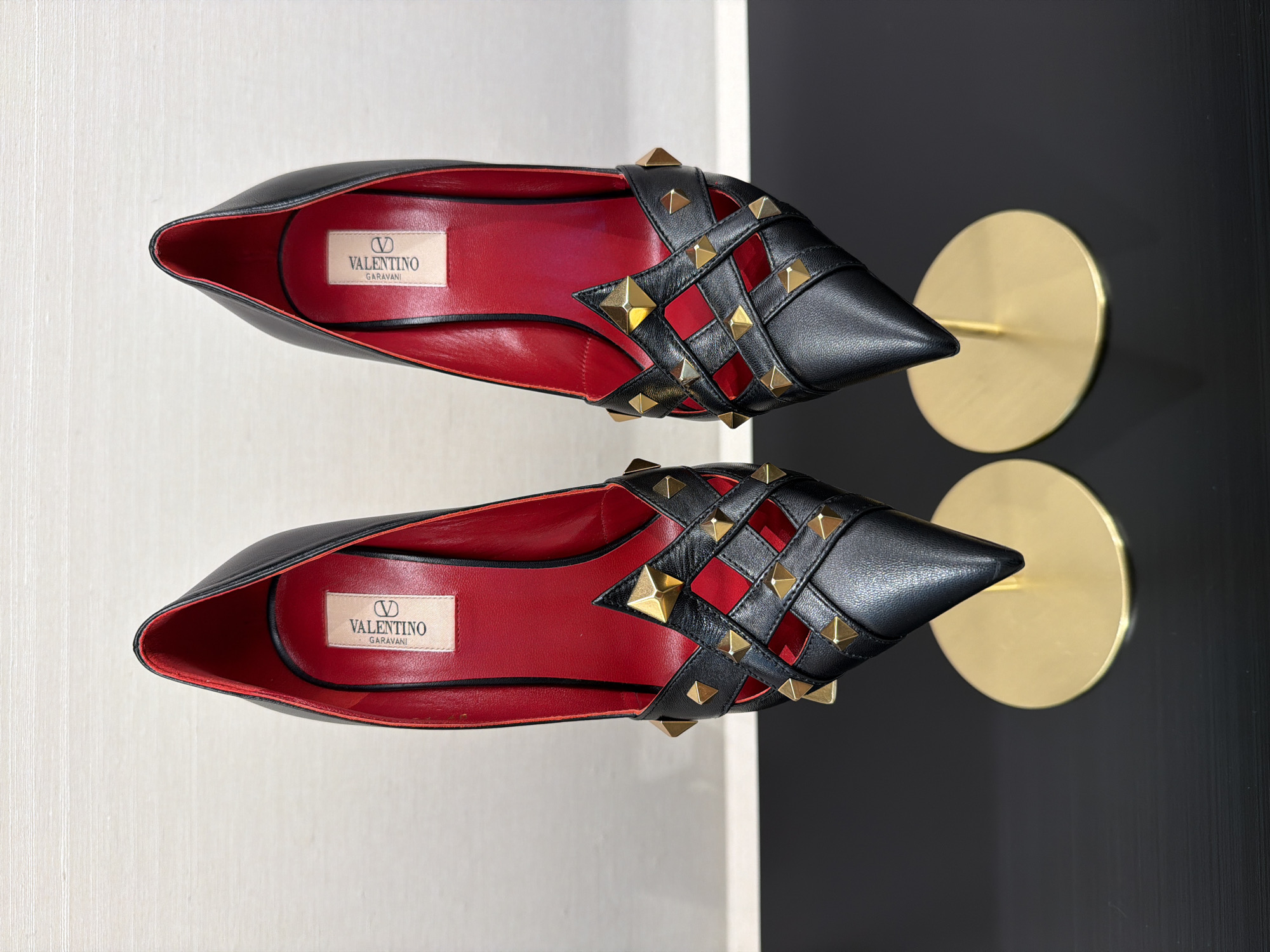 A pair of black Valentino Garavani high-heeled pumps with red interiors are displayed on golden stands against a neutral background. The shoes are made of leather and feature a distinctive woven design on the vamp, adorned with gold pyramid studs. The heels are pointed, and the interior of each shoe displays a label with the Valentino Garavani logo. The image was captured indoors, with the shoes positioned on a dark surface that reflects some light. The lighting is even, highlighting the details of the shoes. The background appears to be a wall with a subtle horizontal texture, suggesting an indoor display setting, potentially in a retail environment or a private collection.