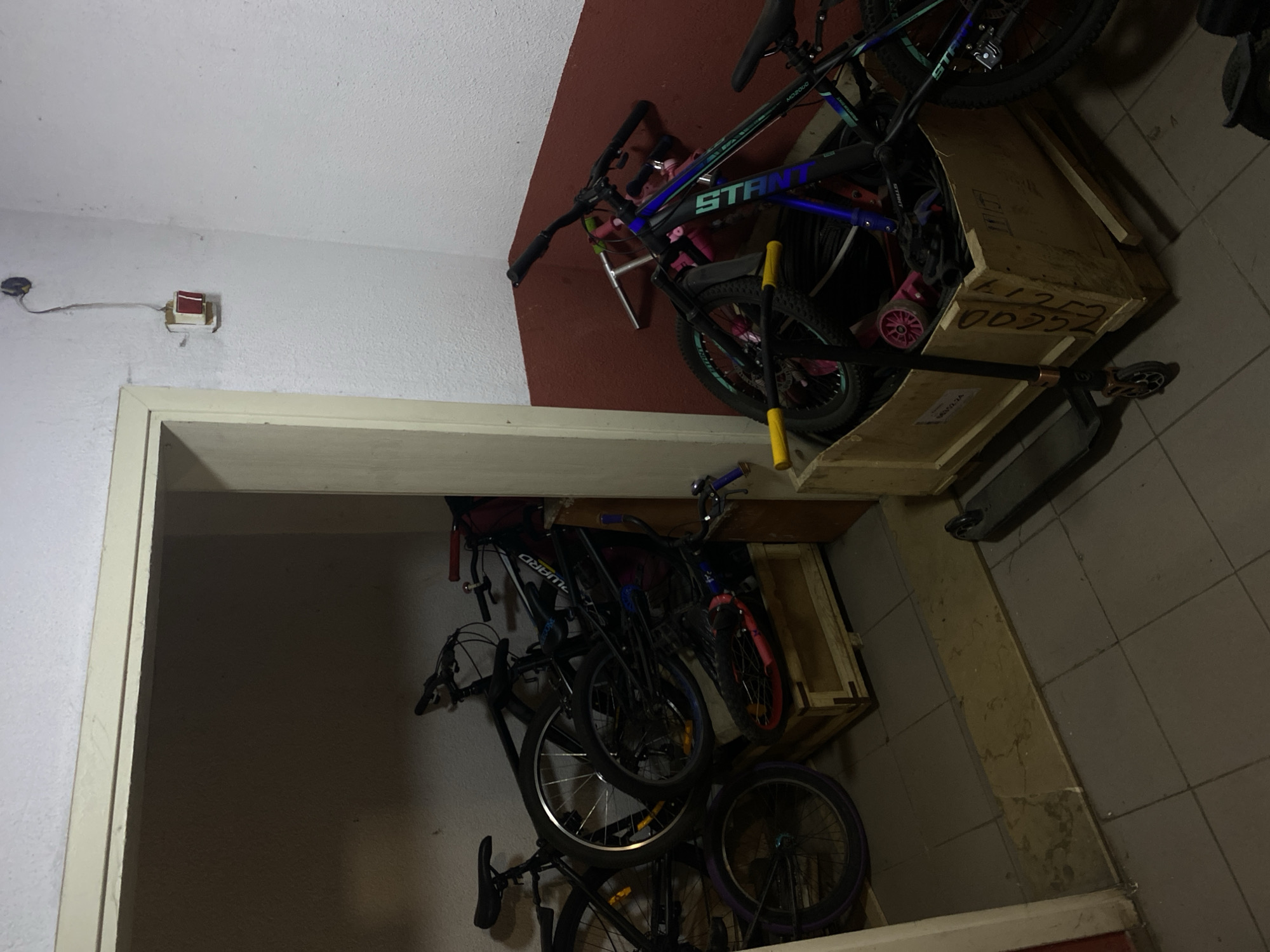 The image shows a storage area in an indoor location, likely a residential building or apartment, in what may be Kazan, Russia. There's a doorway with a cream-colored frame leading into a darker storage room filled with bicycles of varying sizes and colors. The bicycles are stacked somewhat haphazardly. A scooter is next to the bikes. Above the doorway, there's a wall partially painted in a dark red color. The walls appear to be white or cream. On the ceiling near the doorway is a small, possibly electrical, box. The scene is dimly lit, and the focus is on the cluttered storage of bicycles and related items.