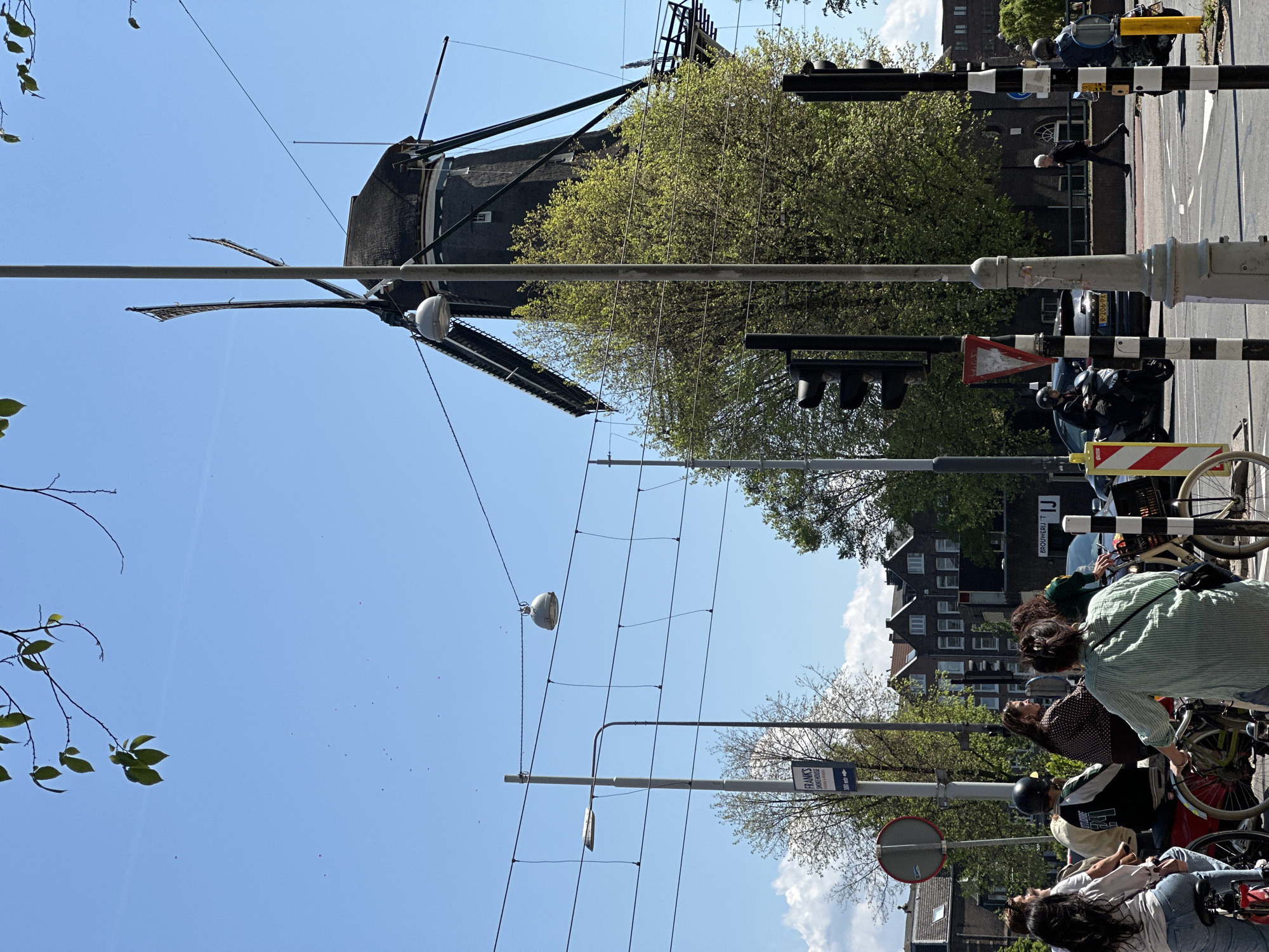 The image shows a street scene in Amsterdam, Netherlands, featuring a windmill in the background. The foreground includes a group of people on bicycles, parked on the side of the road. Overhead are power lines and street lamps. The architecture is typical of Amsterdam, with brick buildings. Traffic lights and street signs are visible along the street. A man is walking in the crosswalk behind a pair of motor bikes.