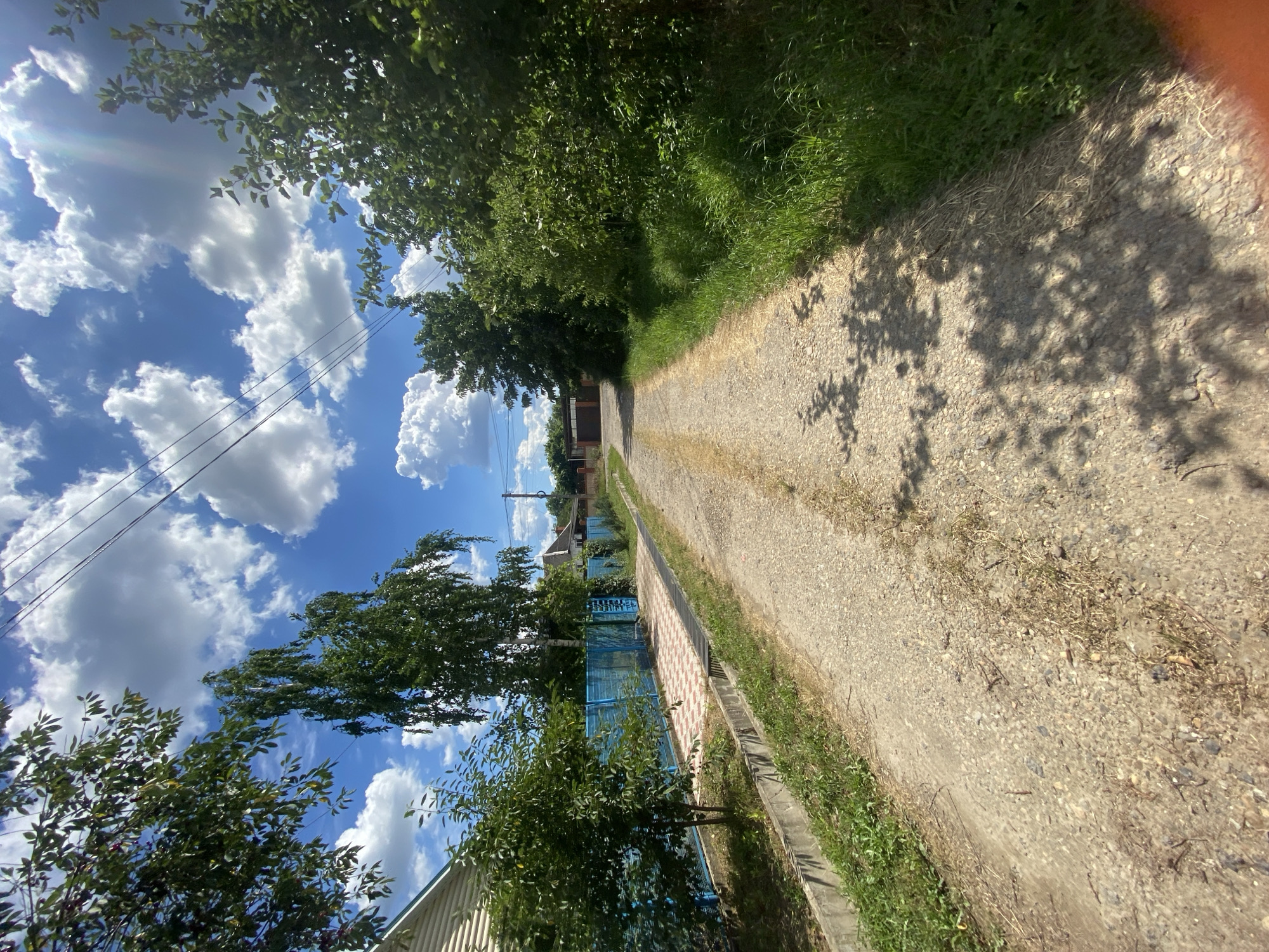 The image depicts a dirt road leading into a residential area in Akhtyrskiy, Russia. The road is lined with vegetation, including trees and grass, and appears to be well-traveled. On the left side of the road, there is a blue fence and a house. On the right side, there are more trees and foliage. In the background, there are houses and a gate. The sky is blue with scattered white clouds, and the sun is shining. The overall impression is one of a quiet, rural setting.