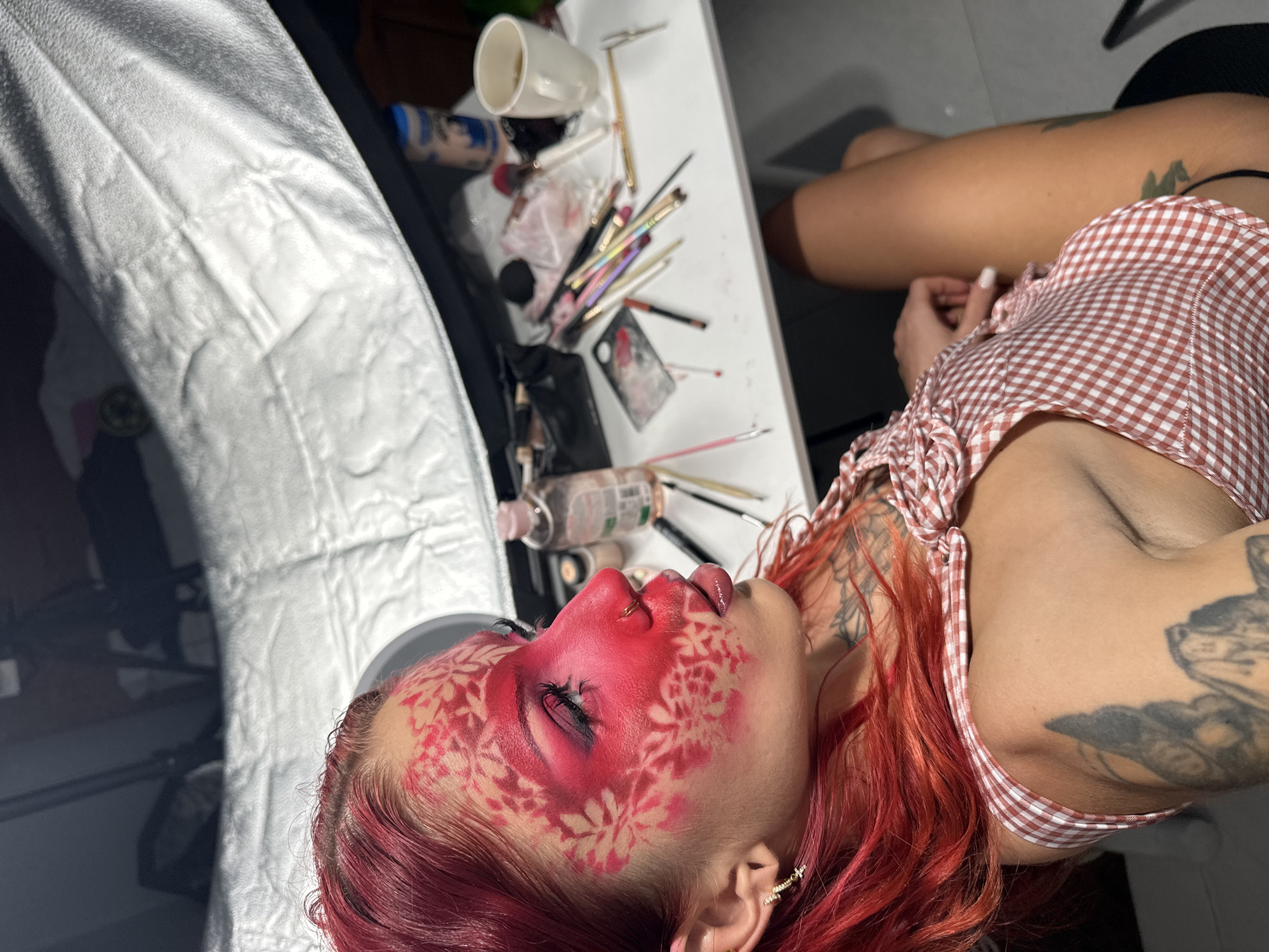 This media file shows a person with vibrant red hair and elaborate makeup. The makeup features a bold red base across the face, with a delicate white floral stencil pattern applied over it, particularly on the cheeks and forehead. The person's eyes are closed, and their lips are slightly parted, suggesting a moment of relaxation or focus during the makeup application. They are wearing a red and white checkered top.
In the background, a table is laden with makeup brushes, bottles of cosmetics, and other beauty supplies, indicating that a makeup session or photoshoot is in progress. A white mug and a white paper covering add to the studio-like setting. Tattoos are visible on the person's arm and shoulder. The lighting suggests an indoor environment, possibly a studio or dressing room. The overall mood is artistic and creative, highlighting the intricate makeup design. The location context provided is North Kuta, Indonesia.