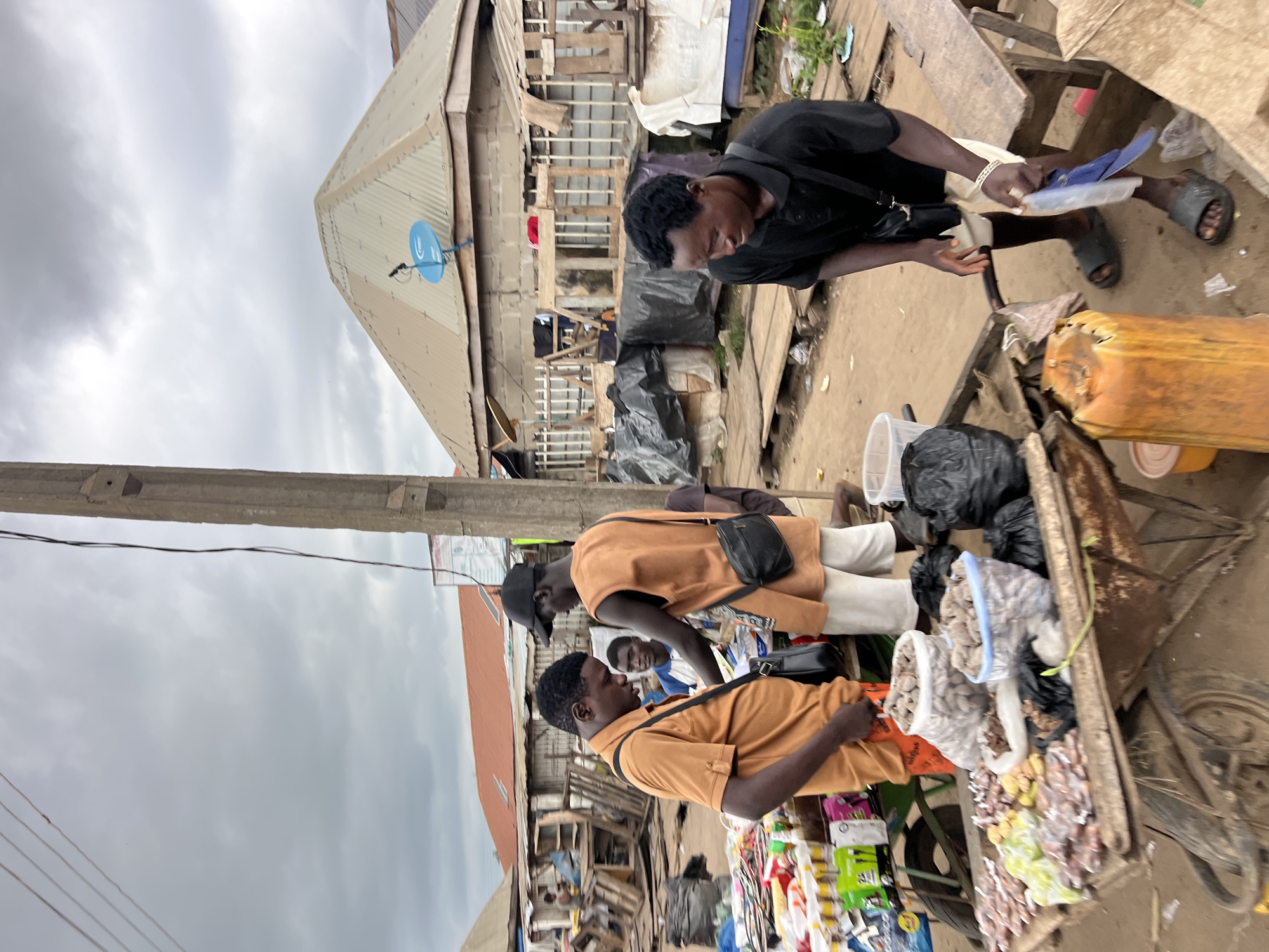 In Oko Amakom, Nigeria, under a cloudy sky, a market scene unfolds. Three men are engaged in commerce around a cart laden with various goods. The man on the left, wearing an orange polo shirt and orange shorts, is bent over the cart, examining items. A black crossbody bag hangs on his shoulder. To his right, a man in an orange sleeveless tunic and white shorts, wearing a black baseball cap, stands and appears to be interacting with the man in orange. He also carries a black crossbody bag. Behind them, another man in a blue and white striped shirt looks on.
The cart itself displays an assortment of products. Two large clear plastic bags filled with small, light-brown items are prominent, as are smaller bags containing similar contents. Various other small items, some wrapped in plastic, are also visible. To the right of the cart, a man in a black t-shirt and white shorts is actively engaged, holding a blue plastic bag and a white object. He wears a bracelet on his left wrist and dark sandals. A large, orange plastic container is situated near his feet.
The environment suggests a bustling outdoor market. Wooden structures and makeshift stalls are visible in the background. Pallets and tarps are used for display and storage. A concrete utility pole with wires runs vertically through the scene, and a satellite dish is mounted on a building's roof. The ground is dirt and debris, indicating a well-trodden market area.