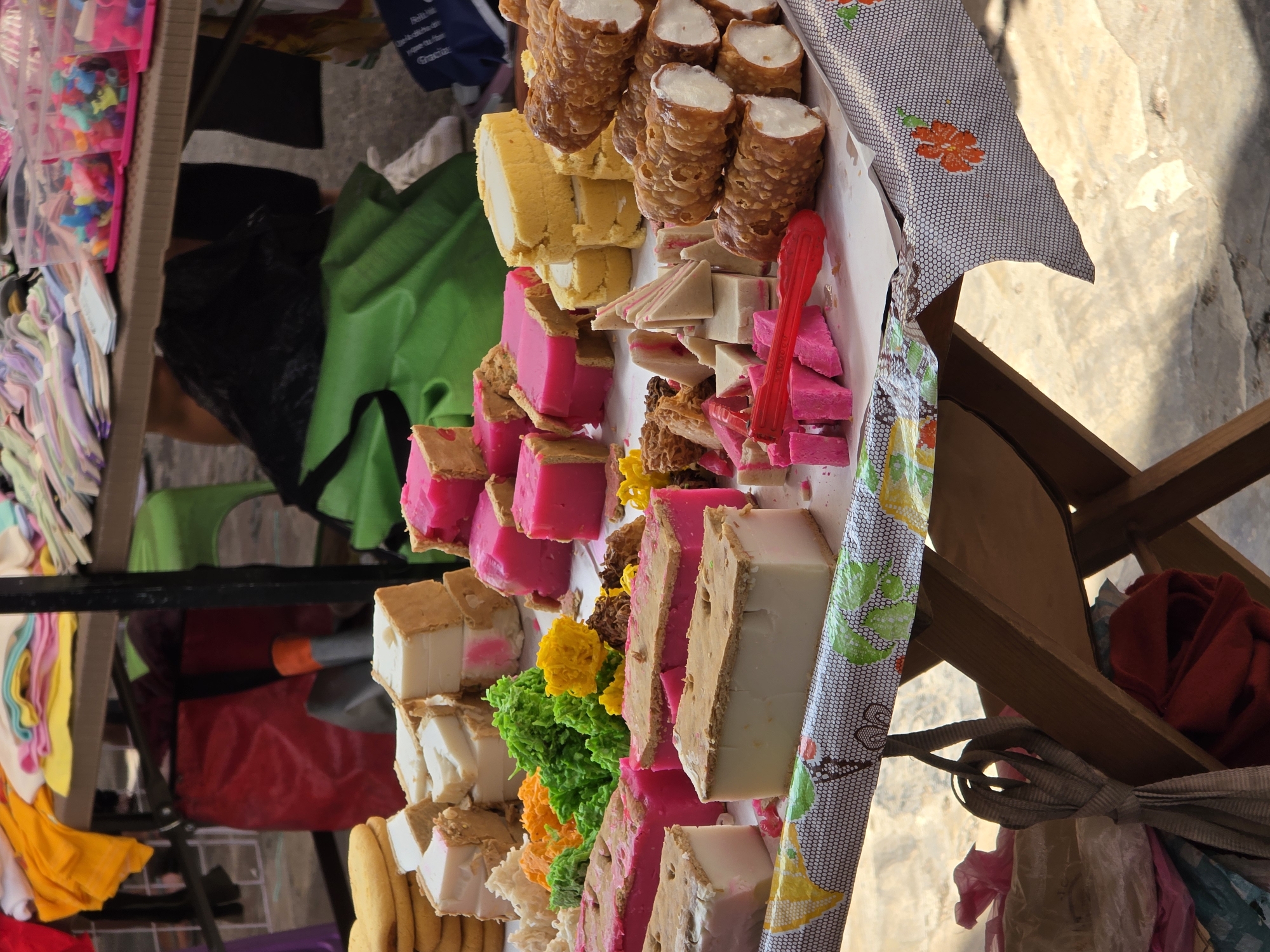This image captures a vibrant market stall in Cuetzalan, Mexico, brimming with an array of traditional sweets and other small goods. The primary focus is a display of confectioneries arranged on a white surface, likely a table or cart, covered by a patterned tablecloth featuring orange flowers and a second, fruit-patterned cover.
In the foreground, numerous sweets are meticulously displayed. These include brown, crispy cylindrical pastries with white filling, resembling cannoli, and slices of yellow cake rolls also filled with a white cream. Dominating the display are bright pink and white rectangular blocks, often with a visible cookie-like base, which could be varieties of *cocadas*, *dulce de leche*, or fruit-based candies. There are also triangular, layered confections combining white and pink sections, alongside smaller, crumbly brown sweets. Decorative elements like clusters of bright green, yellow, and orange sugary candies add to the colorful presentation. A red plastic tong is placed among the sweets, ready for serving.
The background, partially obscured, reveals other market items. To the left, clear plastic bags hold a multitude of small, colorful items, possibly beads, small toys, or accessories. Below these, stacks of folded textiles or clothing in pastel shades are visible. Further back, a green plastic bag and a black plastic bag are seen on the ground or under the stall, with a red bag or cloth item slightly deeper in the scene. The wooden structure of the stall is also discernible.
The scene takes place during the daytime, indicated by the bright, natural lighting and shadows. The setting strongly suggests an outdoor market or street vendor stall, characteristic of Mexican towns. While no people are visible, the neatly arranged goods imply an active selling environment. A blue bag in the background shows partially visible white text, but it is too blurry to decipher definitively.