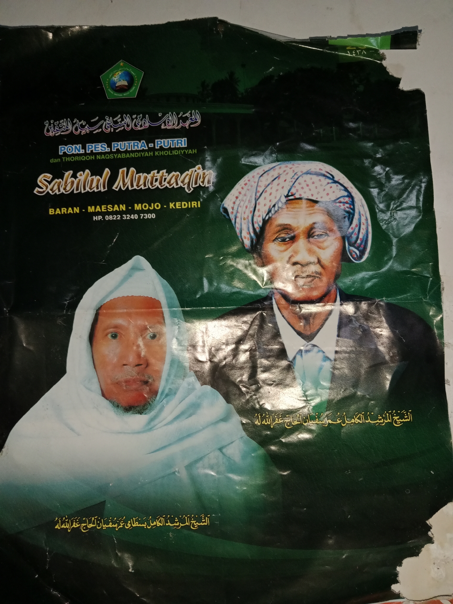 This is a poster featuring two portraits of men, likely religious leaders, against a dark green background. The top of the poster has an emblem with Arabic text and the words "PON. PES. PUTRA - PUTRI" followed by "dan THORIQOH NAQSYABANDIYAH KHOLIDIYYAH" and "Sabilul Muttadin". Below this, it states "BARAN - MAESAN - MOJO - KEDIRI" and a phone number "HP. 0822 3240 7300". This indicates the poster is associated with an Islamic boarding school (Pesantren) in the Kediri region of Indonesia, specifically mentioning the villages of Baran, Maesan, and Mojo, which are located within the Ringinrejo district. The portrait on the left shows a man wearing a white head covering and garment, with a light blue shawl draped around him. The portrait on the right depicts an older man with a patterned headscarf and a suit and tie. Both men are looking directly at the viewer. The bottom of the poster contains Arabic script, which likely identifies the individuals or offers a religious message. The poster appears to be slightly damaged, with some tears and creases, particularly at the top and right edges.