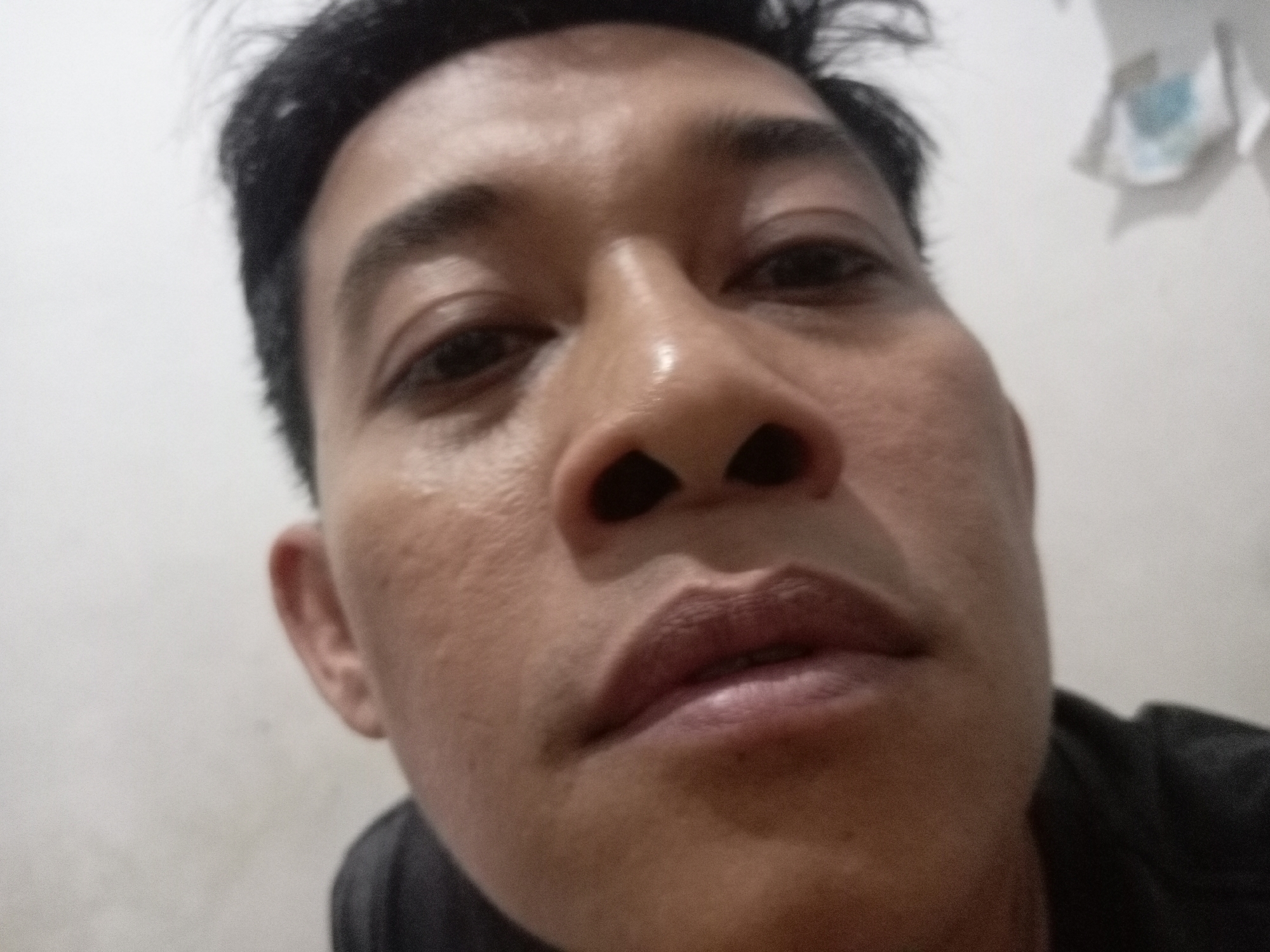 A close-up, selfie-style, low-angle shot of a male individual's face, with a neutral expression, taken indoors. The subject has dark hair and skin with visible pores. The background is a plain, off-white wall with a slightly visible, blurry object on the upper right side. The lighting is diffuse and appears to be from an overhead source, casting subtle shadows. The image quality is moderate, with some graininess and a lack of sharp focus. The location is an unspecified indoor setting in Batam, Indonesia.