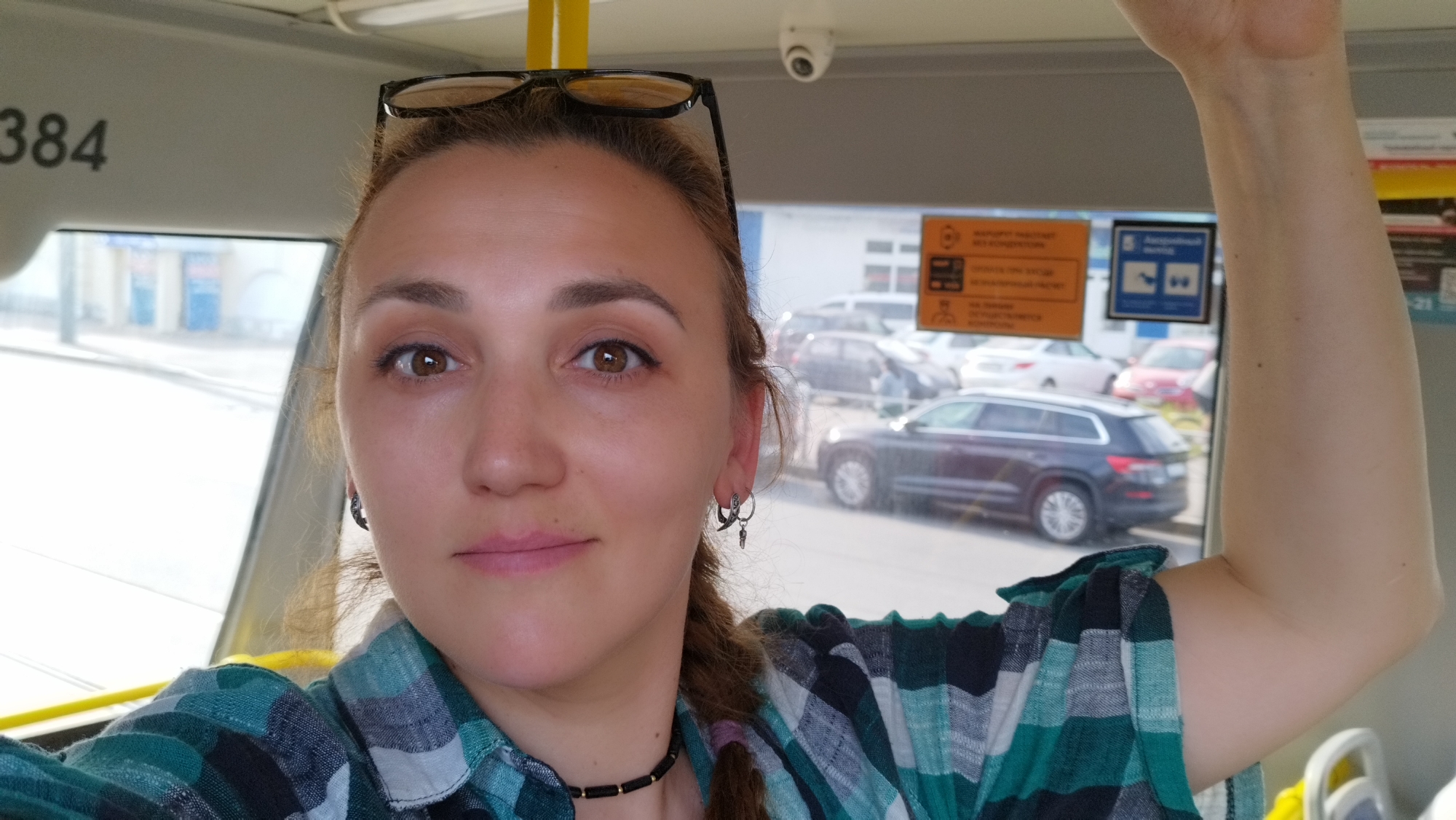 A woman with brown hair is taking a selfie inside a vehicle, possibly a tram, as indicated by the "384" number visible and the yellow pole. She is wearing a green and black plaid shirt and a black necklace. Her glasses are perched on top of her head. She has brown eyes and is looking at the camera with a neutral expression. To the right of her, her arm is raised.
Through the window behind her, a street scene is visible with several cars parked. There are also signs on the window, one of which is orange with Russian text. The setting appears to be a city street during the daytime. The overall mood of the image is casual and candid.