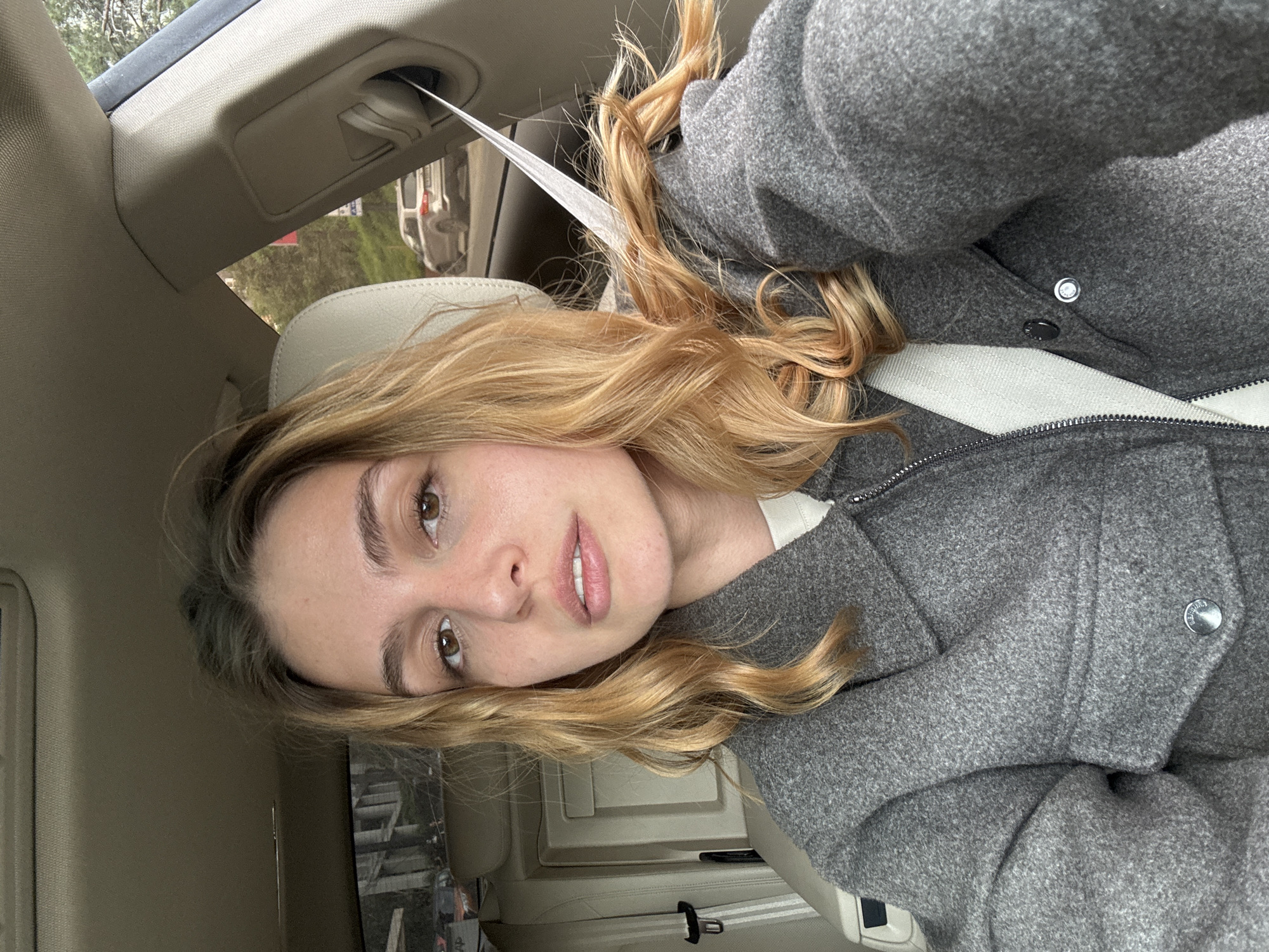 A young woman with blonde curly hair and brown eyes is seated in the driver's seat of a car. She is wearing a gray jacket and a white seatbelt. The interior of the car is beige. Through the car window, trees and a building can be seen. The location appears to be outdoors.