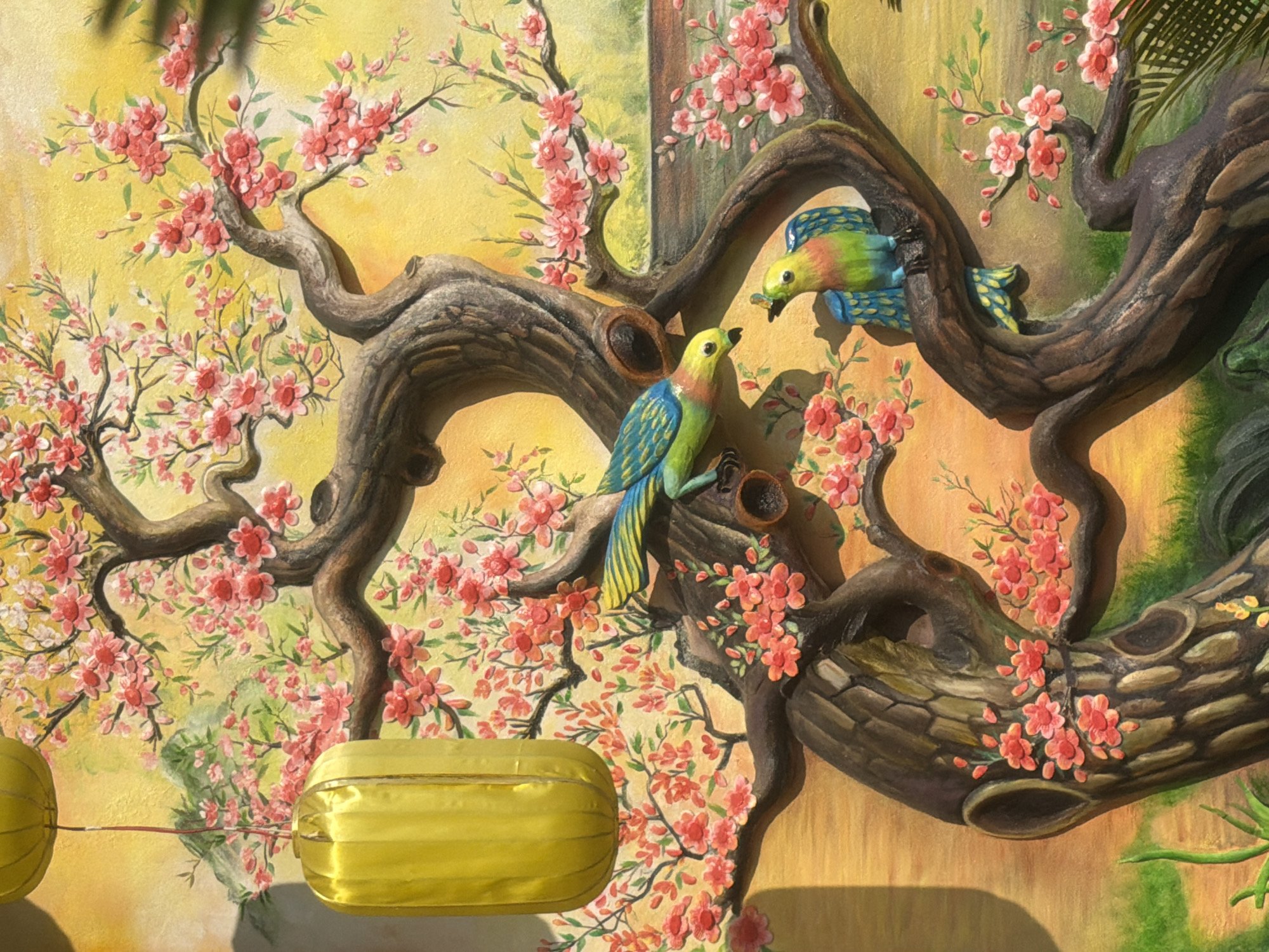 The image depicts a wall mural with a 3D effect in what appears to be a building in Đà Nẵng, Vietnam. The mural features a stylized tree with blooming pink flowers. Two colorful birds are perched on the branches. A yellow, rectangular lantern hangs from the lower left part of the tree. The background is painted in soft yellow hues, creating a bright and serene environment. The mural is detailed and textured, enhancing the visual depth of the scene.