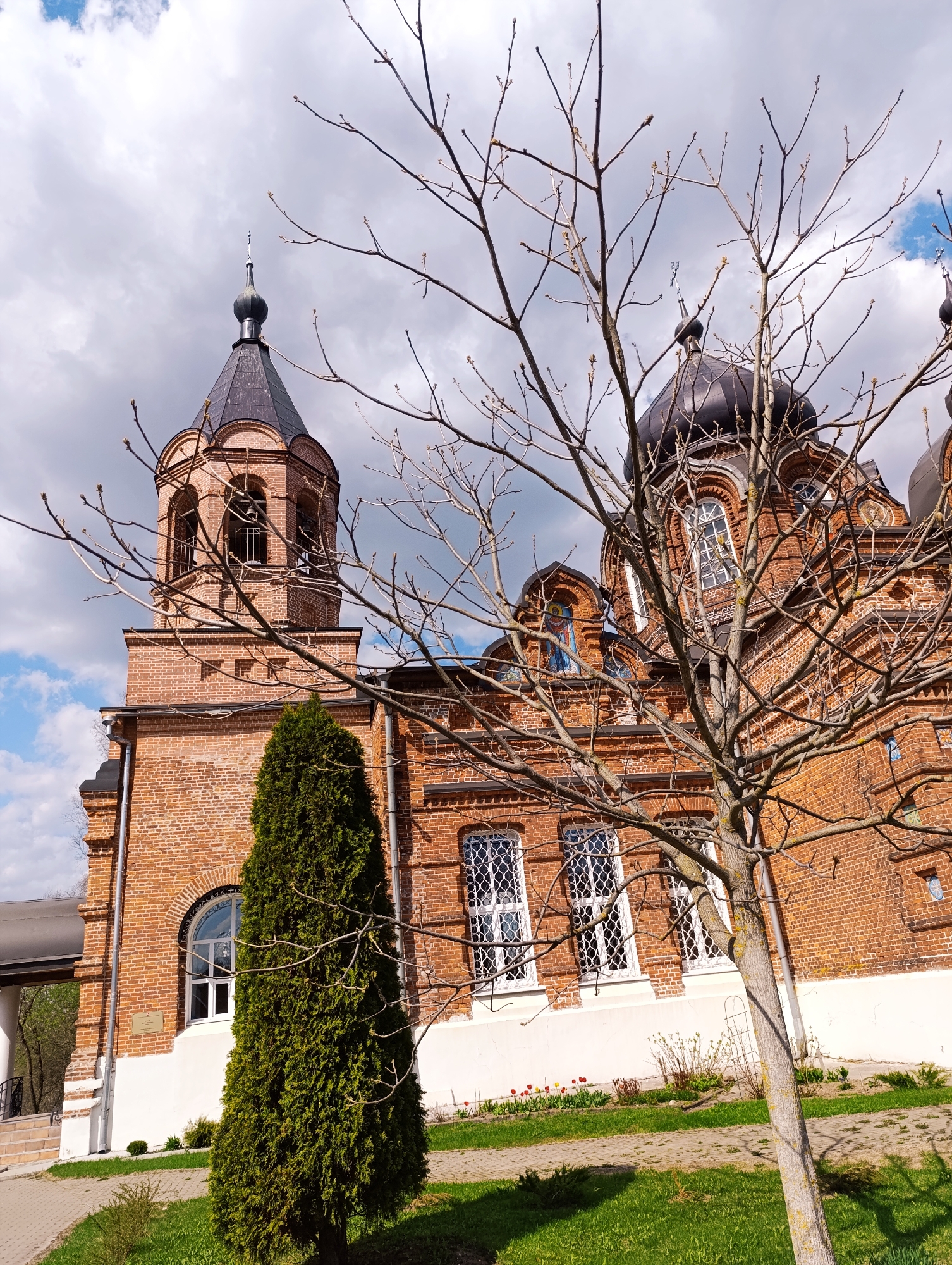 The image captures the Church of the Intercession of the Holy Virgin in Ivanovskoe, Russia. The church is constructed from red brick with white window frames and base. It has several black domes and a bell tower topped with a black spire. In the foreground, there is a bare tree on the right and a green coniferous tree on the left. There is also grass and a paved pathway. The sky is cloudy with patches of blue.