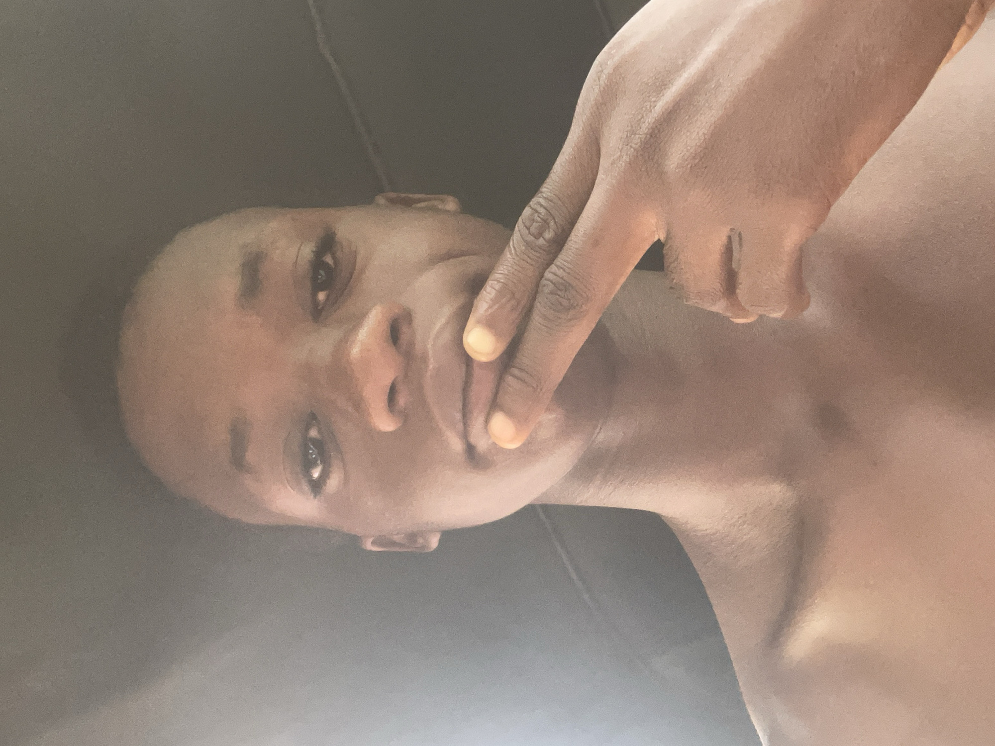 The media file displays an image rotated 90 degrees counter-clockwise. The main subject is a person with dark skin, whose head is positioned on the left side of the frame, looking towards the top edge of the image. The person's body extends horizontally to the right. One hand is raised, with the index finger and thumb gently touching the chin and mouth area. The person's eyes are open and looking forward, conveying a neutral or slightly pensive expression. The upper body, including shoulders and chest, is visible and appears shirtless or wearing very minimal clothing. The lighting is soft and even. The background is a plain, dark, unidentifiable surface, occupying the top and bottom portions of the frame. There are no discernible objects, text, or specific location cues visible within the image. The location is provided as Nkpor, Nigeria.