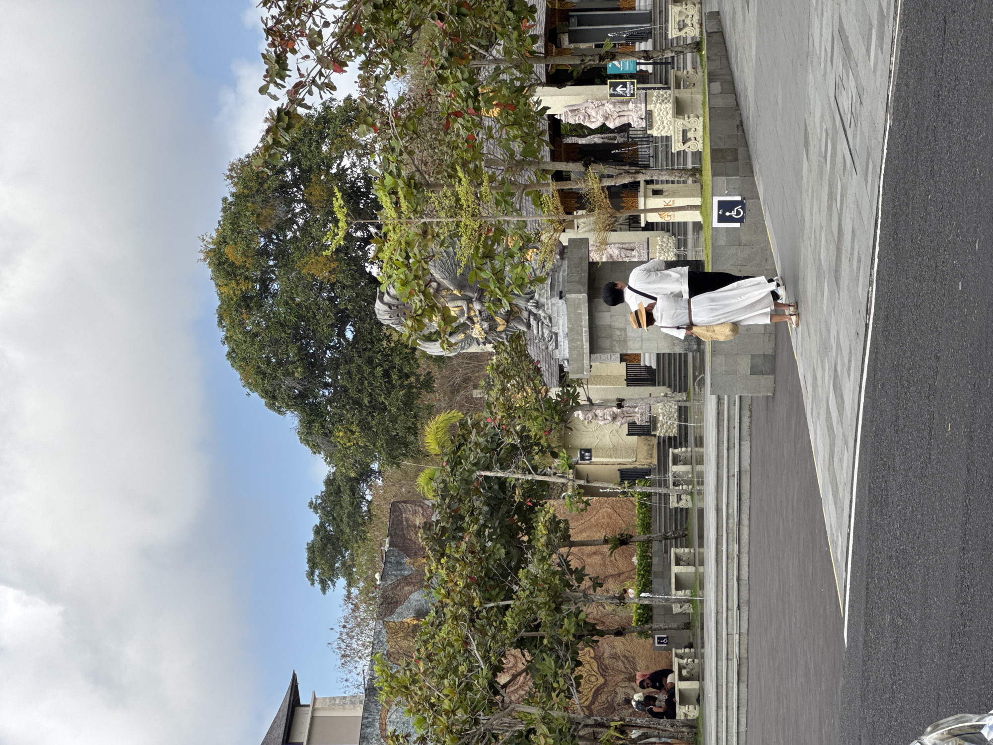 In Kuta Selatan, Indonesia, a paved area shows a couple walking away from the camera, a woman in a white dress and straw hat, and a man in a white shirt and black pants. They are on a tiled walkway, with a dark asphalt road to their left. Behind them, a large stone statue of a winged creature with multiple arms is visible, part of a larger architectural complex. Numerous trees with green foliage are scattered throughout the scene, some bearing red leaves or small red fruits. In the background, a building with intricate carvings and several smaller statues are present. People are seated on benches near a water fountain. The sky is overcast with clouds.