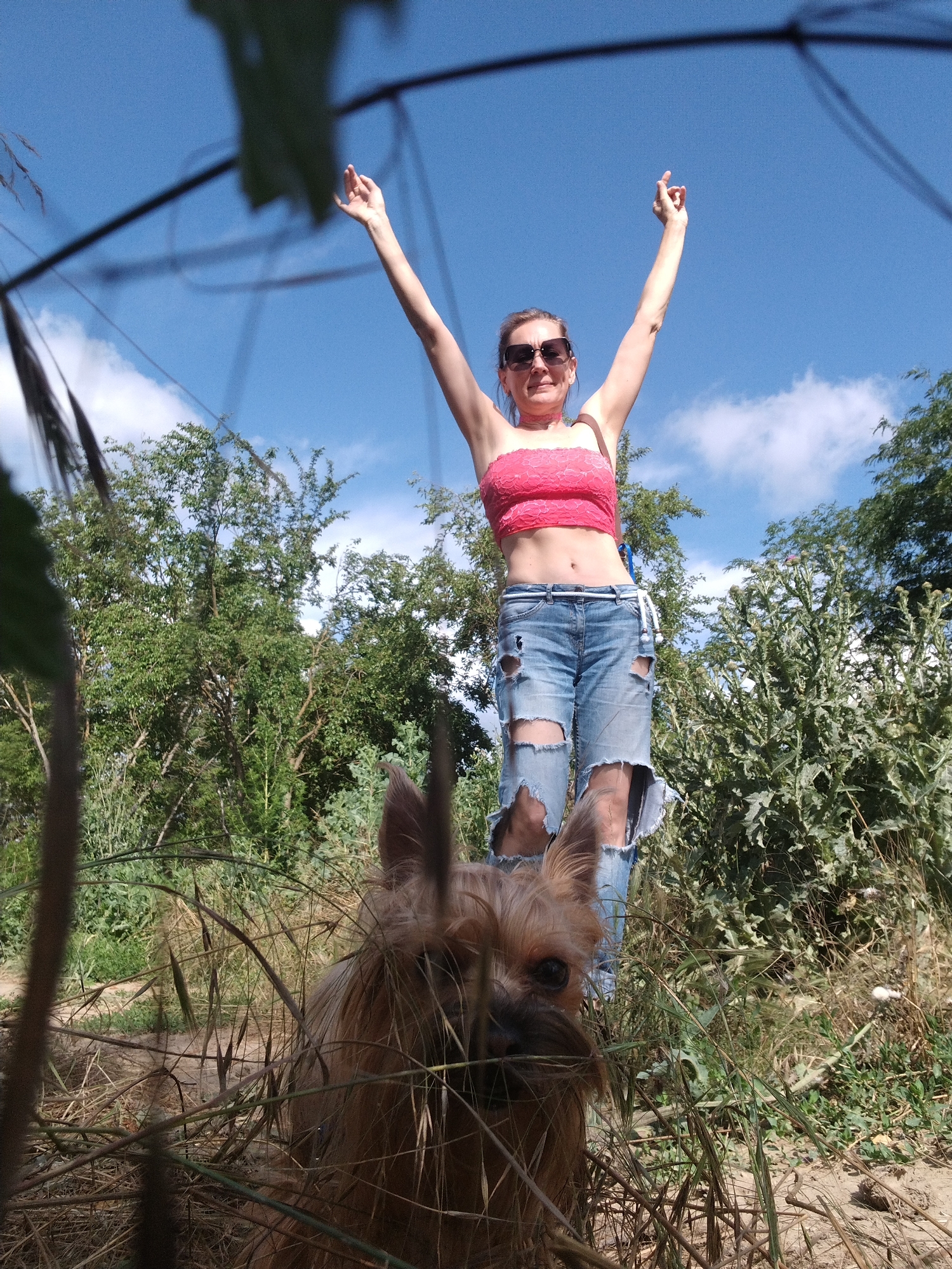 A medium shot captures a woman standing in a natural, outdoor setting, likely near Fyodorovskaya, Russia. She is dressed in ripped denim jeans and a pink lacy crop top, with her arms raised in the air. She wears sunglasses and a choker necklace. In the foreground, a small, tan dog is visible, partially obscured by tall grass. The background consists of lush green trees and a clear blue sky with scattered clouds. The overall impression is one of a casual, outdoor activity.