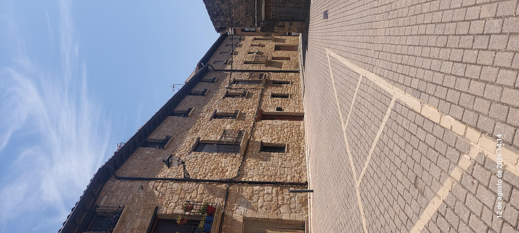 The image depicts a sun-drenched plaza with cobblestone paving and a row of traditional stone buildings lining one side. The architecture features rustic stone walls, dark-shuttered windows, and balconies with wrought-iron railings. A street lamp extends from one building, and potted plants are visible on some balconies. The ground is marked with painted lines, suggesting designated parking spaces. In the background, a narrow street leads away, with another stone building visible on the right. The sky is clear blue with wispy white clouds. The overall scene conveys a sense of an old European town square. A timestamp in the lower left corner indicates "12 de agosto de 2026 14:14."