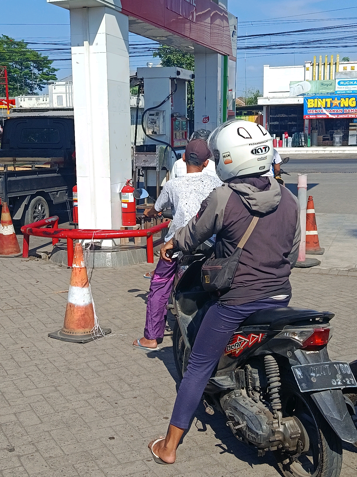 In Tempeh Lor, Indonesia, a bright, sunny day unfolds at a gas station. Two people are on a motorcycle, with the rider in front wearing a white helmet adorned with the "KYT" logo and a sticker that reads "#ASMARAKACAU". They are dressed in a dark grey hooded jacket and dark blue pants, with a black bag slung across their shoulder. The passenger behind them wears a patterned white shirt and purple pants, with a brown baseball cap. The motorcycle, a black Honda Beat, has a license plate that reads "N 4247 YBD 02-27". In the background, a black truck is parked, and a red fire extinguisher stands near a gas pump. Orange traffic cones with white tape are placed around the area, one with white string tied to it. The ground is paved with grey interlocking bricks. A sign for "BINTANG" is visible on a building in the distance, advertising "MENYEEDAKAN BAHAN BAKAR".