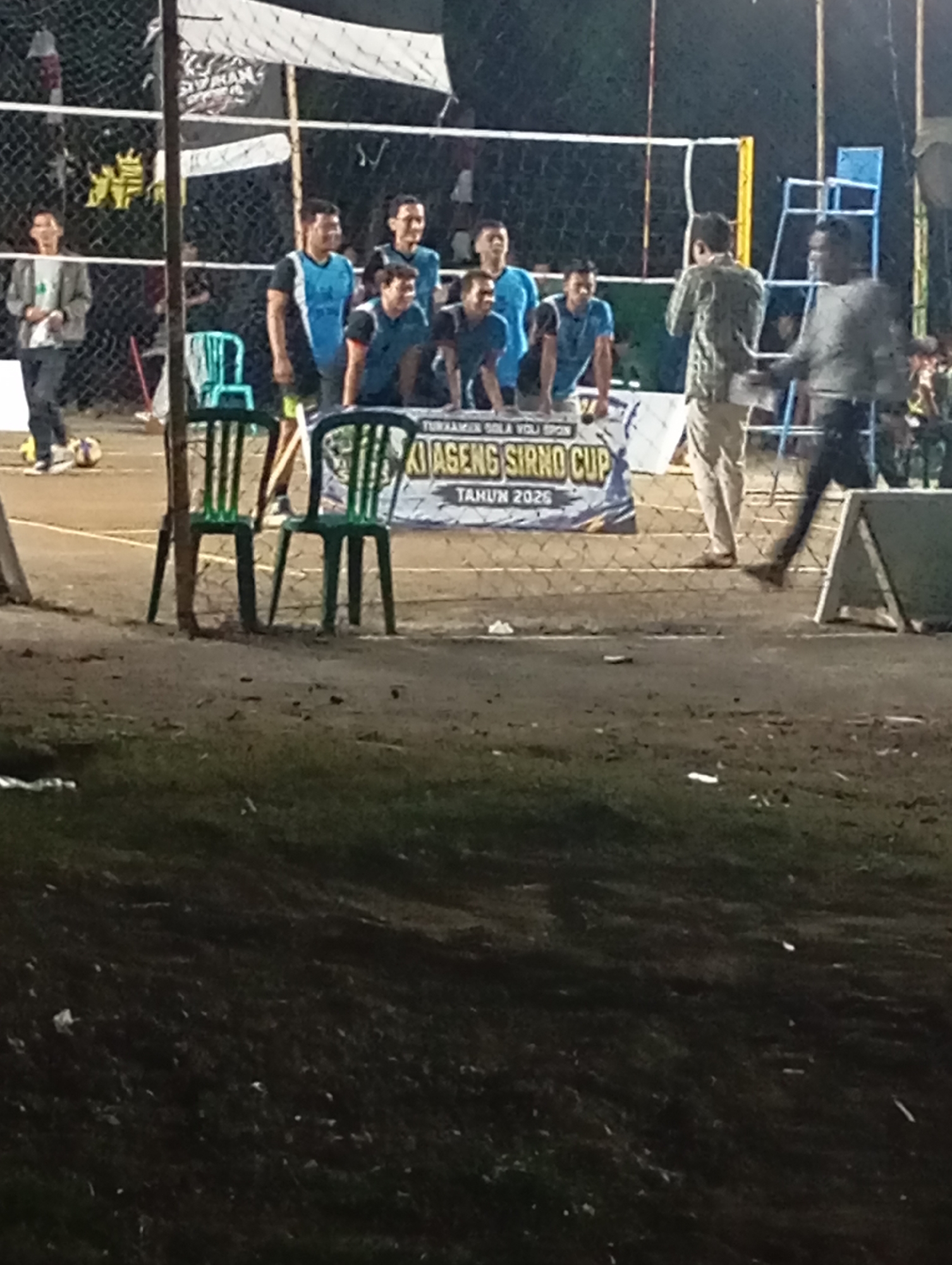 The image depicts an outdoor volleyball court at night in Pacitan, Indonesia. Several young men in athletic wear are visible on and around the court, some appearing to be players and others observers. A net stretches across the court, and a banner with Indonesian text, including "AGENG SIRNO CUP TAHUN 2026," indicates an upcoming or ongoing tournament. In the foreground, two green plastic chairs are placed on the ground. The lighting suggests an event taking place in the evening or night. The overall scene captures a moment of sporting activity in a community setting.