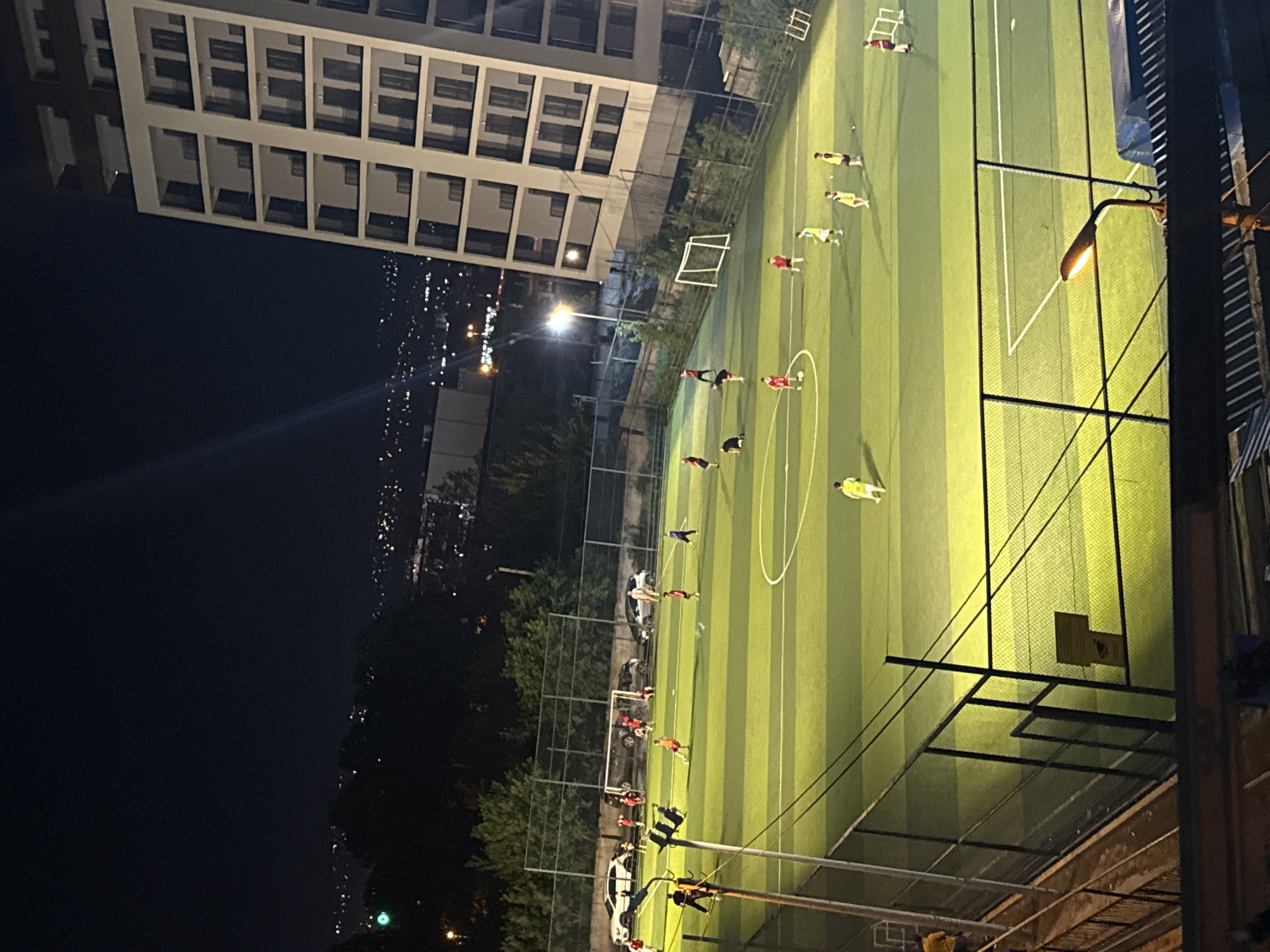 An aerial nighttime shot captures a soccer game on an illuminated artificial turf field in Tbilisi, Georgia. A modern, multi-story white building with numerous balconies dominates the right side of the frame, casting shadows over a portion of the field. The cityscape is visible in the background, with scattered lights indicating urban development. On the field, several individuals, dressed in red and yellow soccer attire, are engaged in a match. Some players are actively running, while others are positioned on the field. A white soccer goal is visible on the field. Cars are parked near the edge of the field, and a fence encloses the playing area. The lighting from the field's lamps creates a bright, green glow on the turf, contrasting with the dark night sky.