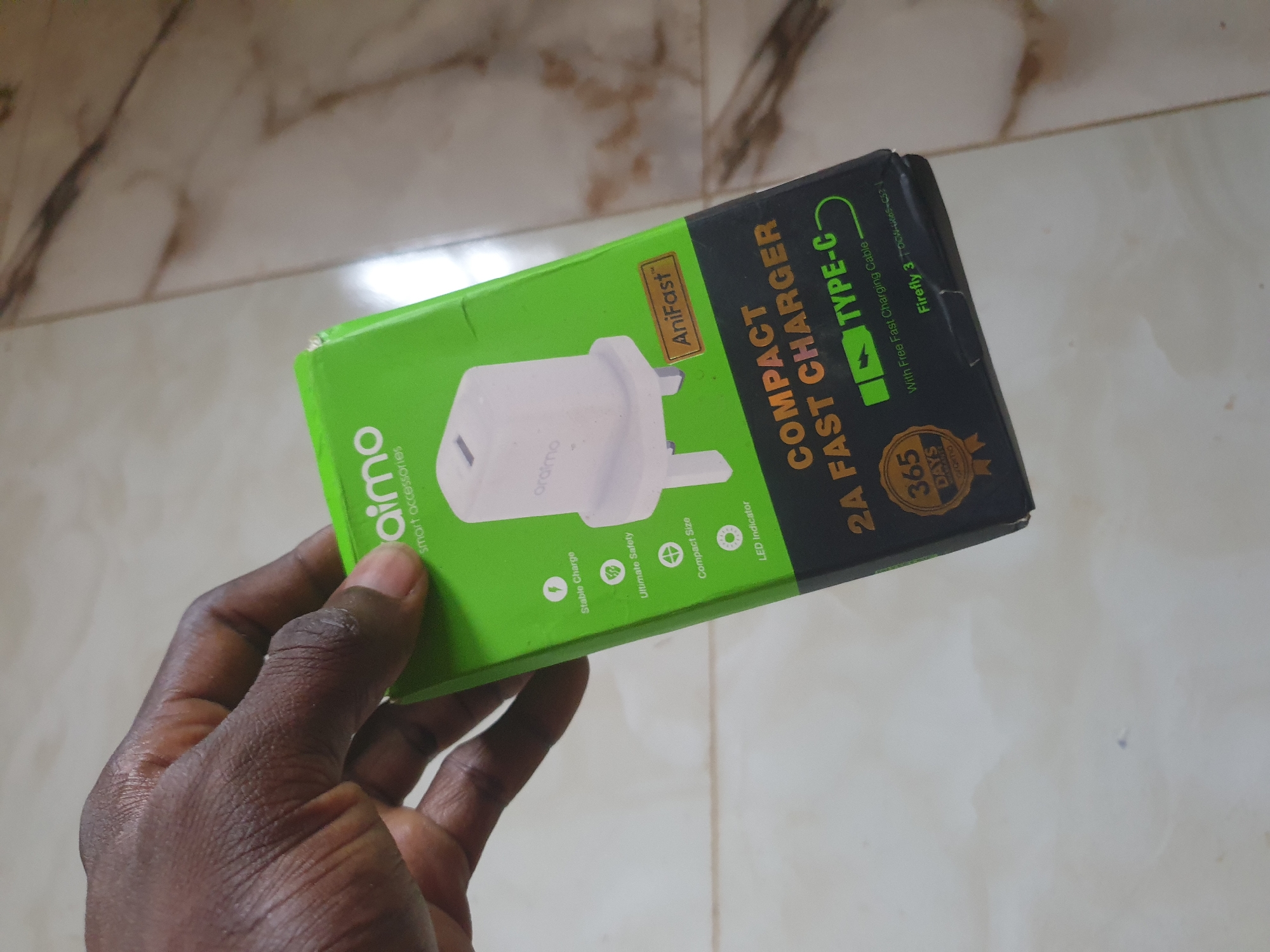 A hand is holding a green and black box that contains a white Orimo smart charger. The box has text on it, including "Orimo smart accessories," "AniFast," "COMPACT FAST CHARGER," "TYPE-C," and "365 DAYS WARRANTY." The charger itself is white and has a single port. The background is a light-colored, tiled floor.