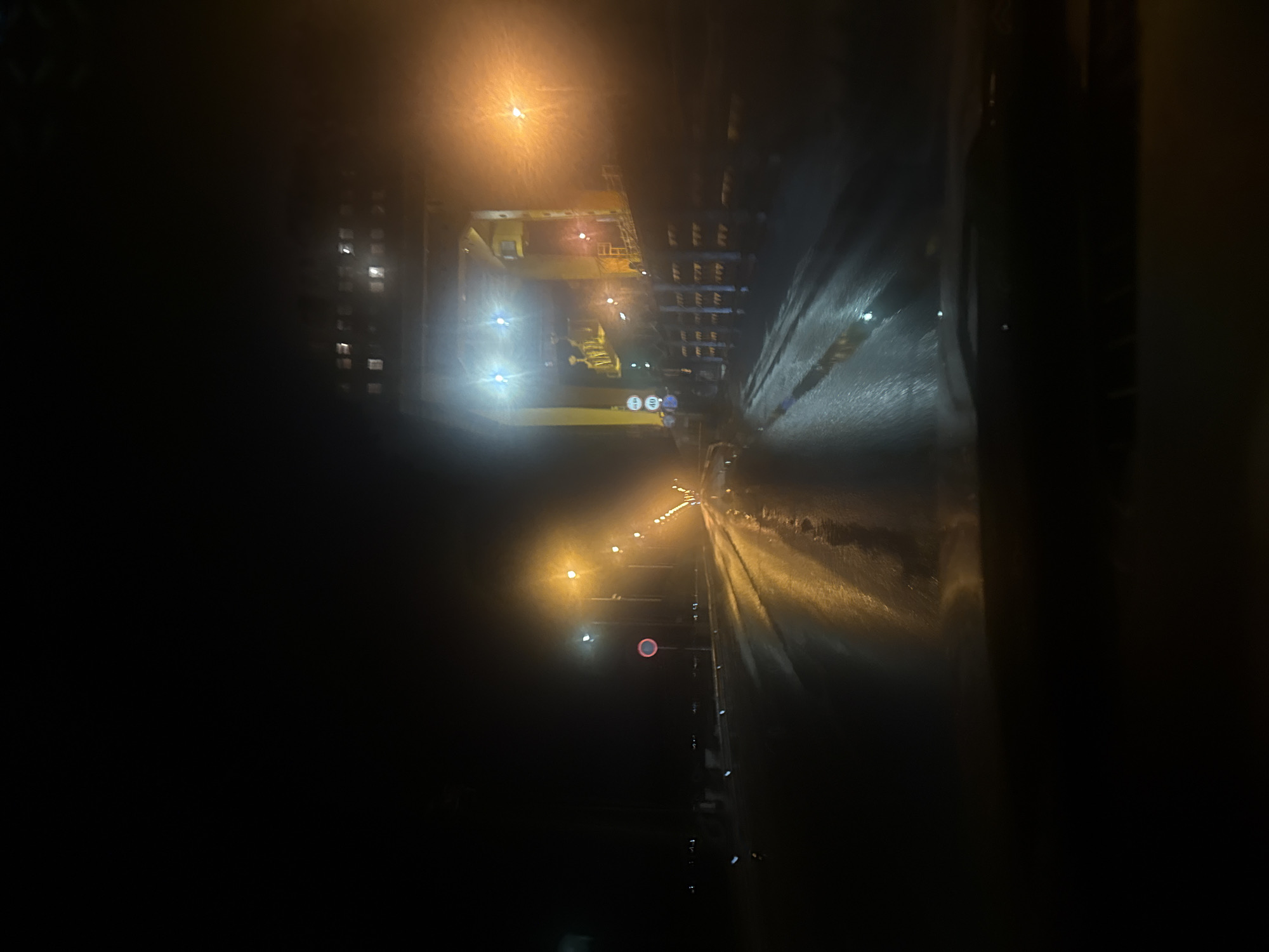 The image depicts a night scene in Chaikovsky, Russia, showing a road with streetlights and a large industrial structure overhead. The road appears wet, reflecting the light. A building is visible on the left with illuminated windows. Traffic signs are present, including a "no entry" sign. The industrial structure appears to be a gantry crane or similar equipment.