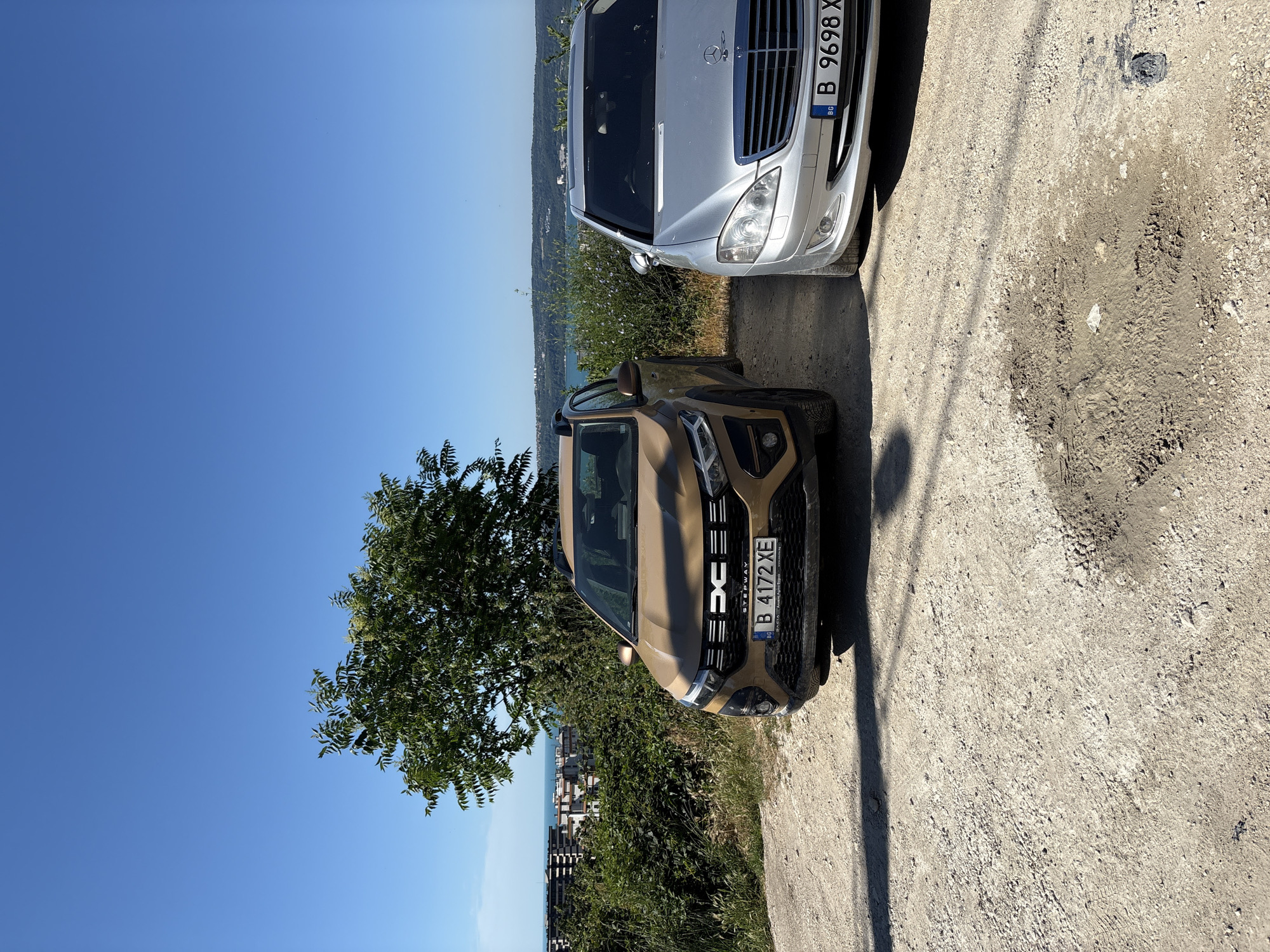 The image depicts a bright, sunny day in Varna, Bulgaria, with a clear blue sky overhead. Two cars are parked on a gravelly surface on a hillside overlooking the sea. The car in the foreground is a tan Dacia Stepway, its front bumper and license plate, which reads "B 4172 XE," clearly visible. To its right is a silver Mercedes-Benz S-Class, also parked, with its license plate "B 9698 X" partially obscured.
Behind the cars, lush green vegetation covers the hillside. In the distance, the city of Varna is visible, with apartment buildings and structures nestled amongst the greenery, leading down to the blue expanse of the Black Sea. The scene suggests a scenic overlook, possibly a parking area on the outskirts of the city, offering a panoramic view of the coastal landscape. No people are present in the image, and the cars appear to be stationary. The ground is uneven and a patch of mud is visible in the lower right section of the image.