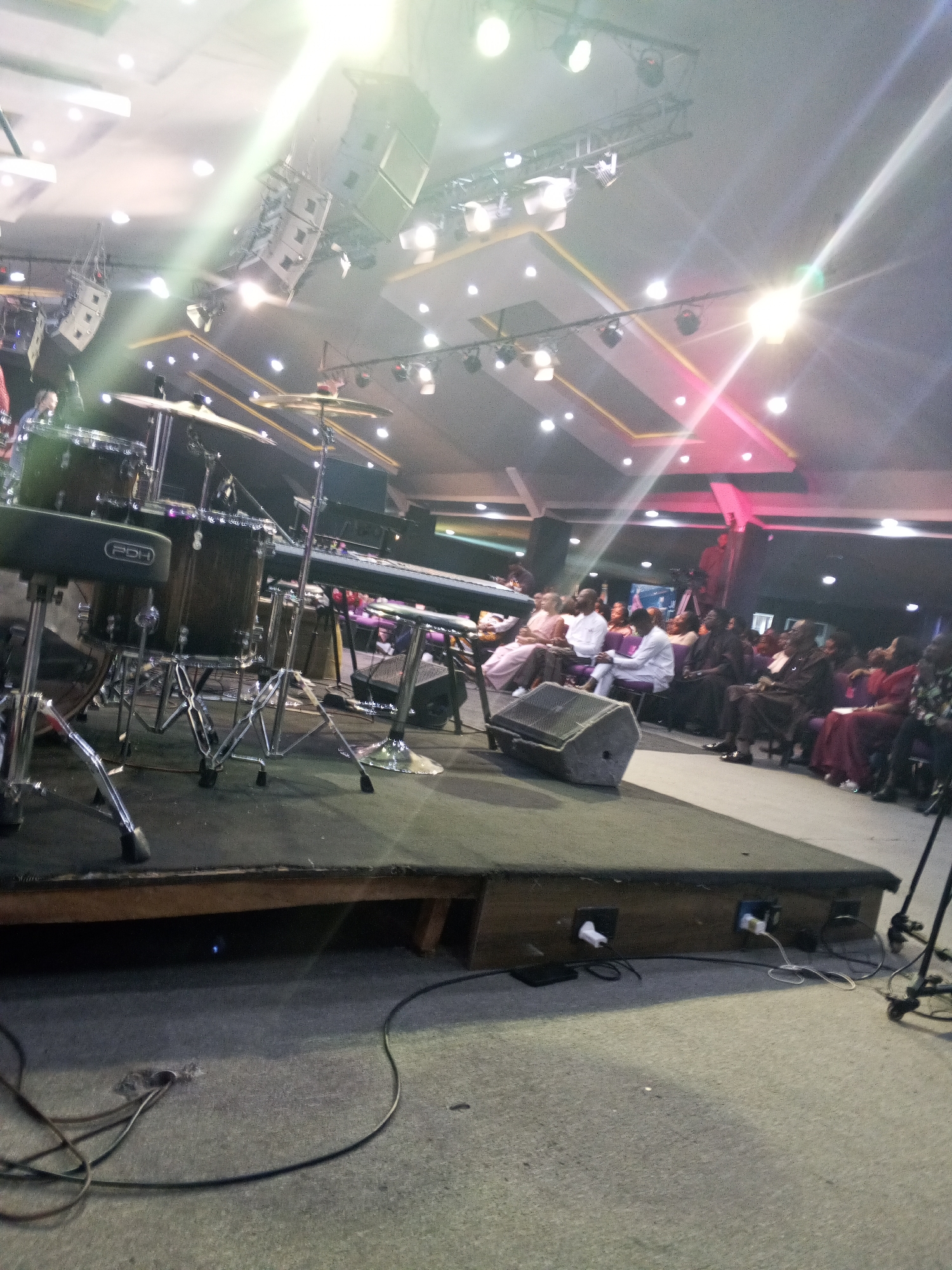 The image captures an indoor event setting, likely a performance or gathering, situated in Abuja, Nigeria. The perspective is from a low vantage point on the floor, looking across the front of a stage towards a seated audience.
The stage is a dark, elevated platform, possibly covered in dark carpeting. On the left, a five-piece drum set is visible, including a bass drum, snare drum, various cymbals (hi-hat, crash/ride), and a drum stool branded "PDH." To the right of the drums, a black electronic keyboard is set up on a stand. A dark gray floor monitor speaker is positioned on the stage, facing the viewer. The front face of the stage structure reveals electrical outlets with white plugs connected, and black and white cables are coiled on the concrete floor in front of the stage. A faint blue light is visible emanating from beneath the stage.
Above the stage, multiple professional stage lights and large, light-colored line array speaker cabinets are suspended from metal trusses. The ceiling is an elaborate multi-layered structure featuring recessed panels, white surfaces, and gold-colored trim, illuminated by numerous circular downlights and spotlights. Intense light sources create prominent lens flares across the upper half of the image, with some lights casting a white or yellow glow and others, further back, emitting a reddish-magenta hue.
Beyond the stage, a seated audience is arranged in rows of chairs, facing forward. The attendees appear to be adults, predominantly of African descent, dressed in various attires including formal and semi-formal wear such as white shirts, light pink dresses, dark suits, and red garments. They are observing the stage. A video camera on a tripod is faintly visible in the far-right background, positioned among the audience. The background walls appear dark.