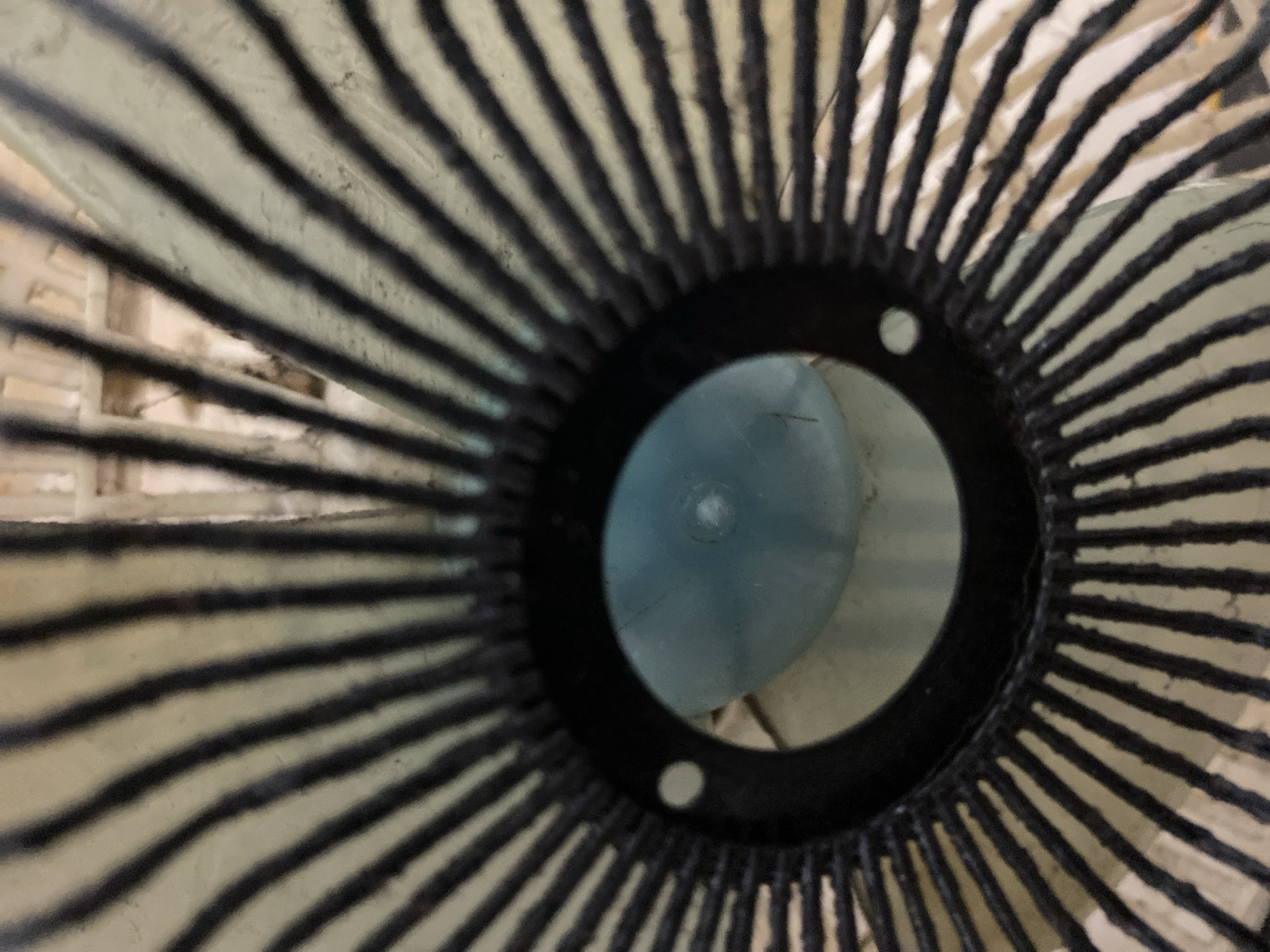 The image shows a close-up, low-angle view of a fan guard. The fan guard is black with thin, evenly spaced black bars radiating outwards from a central black ring. Through the center of the ring, a rounded, light blue object is visible, possibly the motor housing or a light. The background is blurred but appears to be an interior space with wooden slats or beams. The image is undated and does not contain any specific information about its location in Banda Aceh, Indonesia. There are no people or discernible actions depicted in the image.
