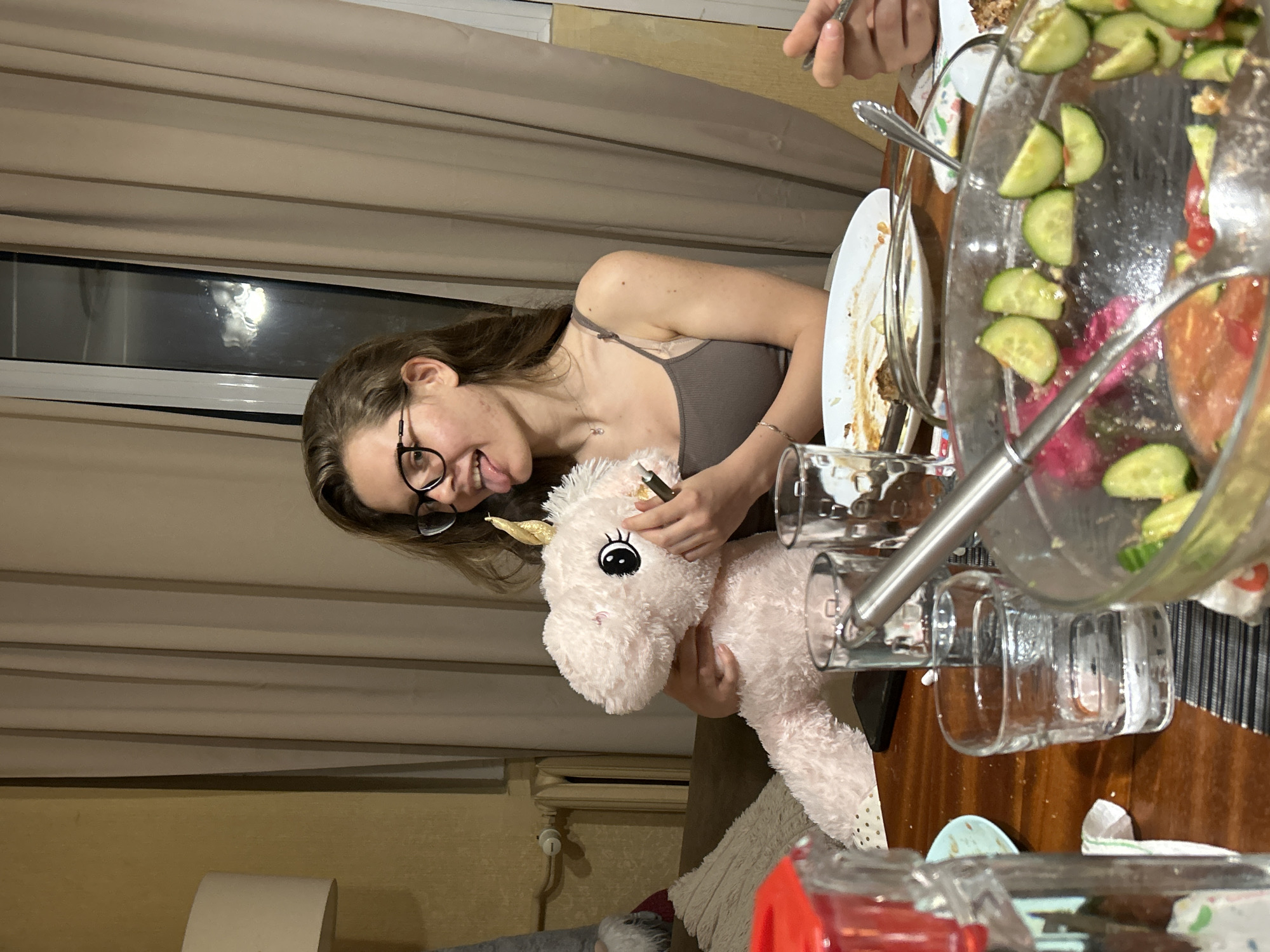 In Sofia, Bulgaria, a young woman with brown hair and glasses is sitting at a wooden table indoors. She is holding a light pink stuffed unicorn in her arms and sticking her tongue out. There are several plates with leftover food, glasses of water, and a salad bowl on the table. The background shows beige curtains and a radiator, suggesting a domestic setting.