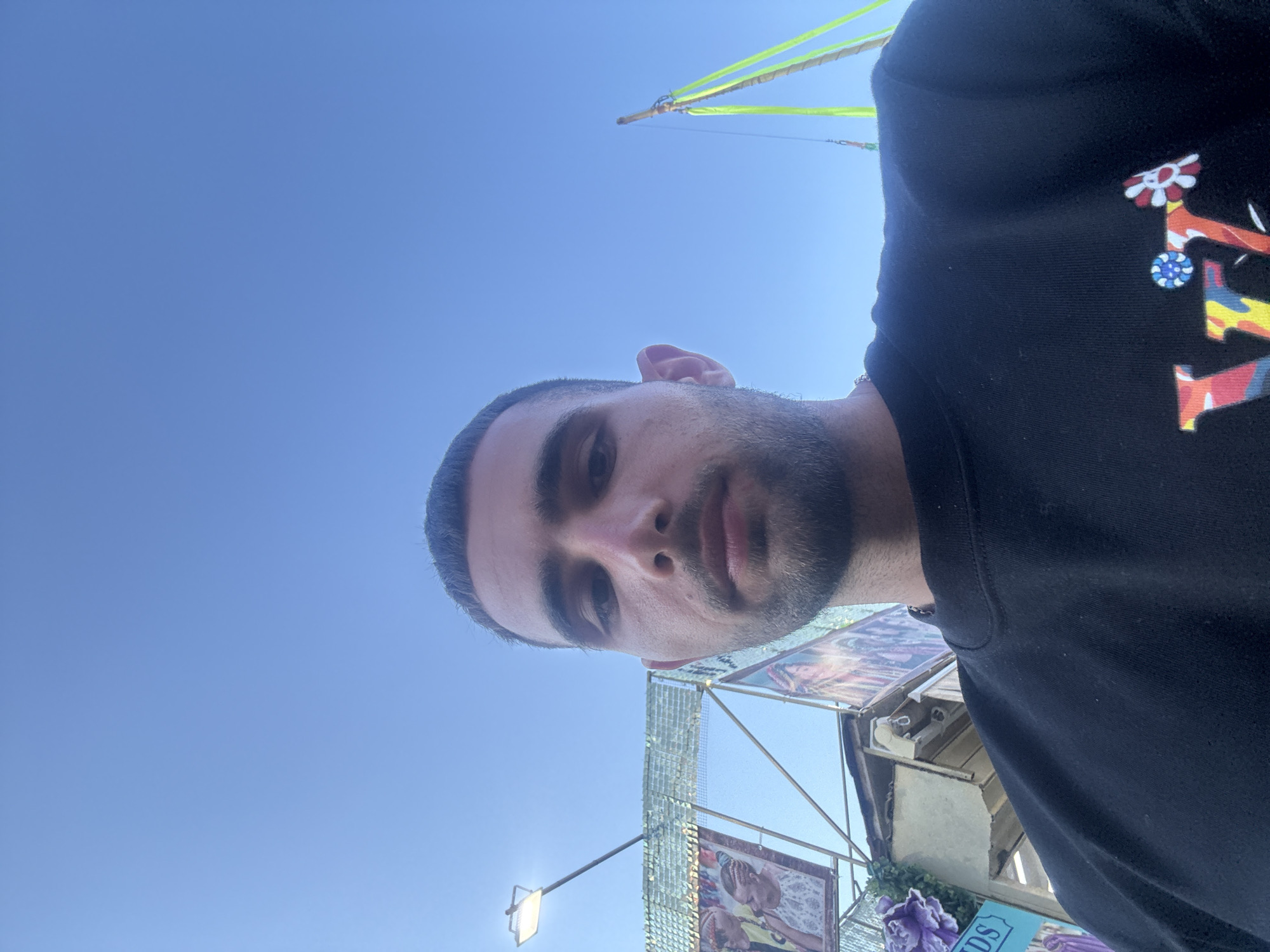 In Nesebar, Bulgaria, a close-up selfie captures a young man against a clear blue sky. He has short dark hair and a light beard. He is wearing a black sweatshirt with a colorful graphic on the front. In the background, a bright lamp is visible, along with scaffolding and banners displaying images of people. The scene appears to be outdoors, possibly at an event or fair, given the festive banners and structures.