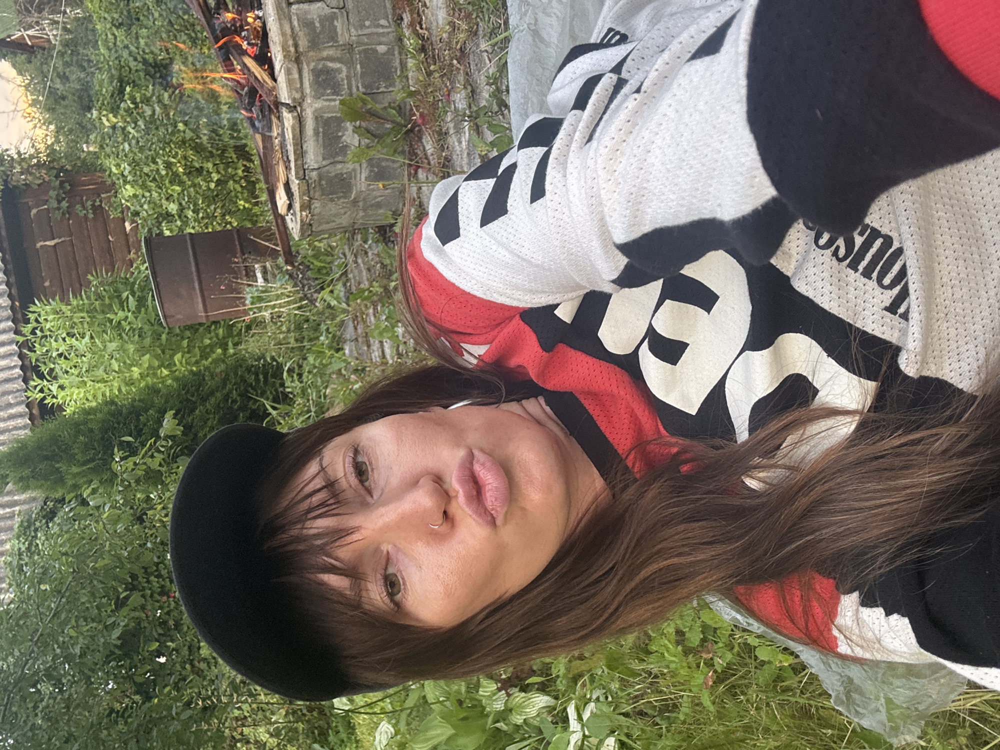 A woman with long brown hair and bangs, wearing a black baseball cap and a red, black, and white long-sleeved shirt, poses for a selfie outdoors. She has a nose ring and is puckering her lips. The background shows a fire pit with flames, a rusty barrel, and lush green foliage, suggesting a rural or suburban setting in Krasnaya Presnya SNT, Russia. A corrugated metal roof structure is visible in the upper background.