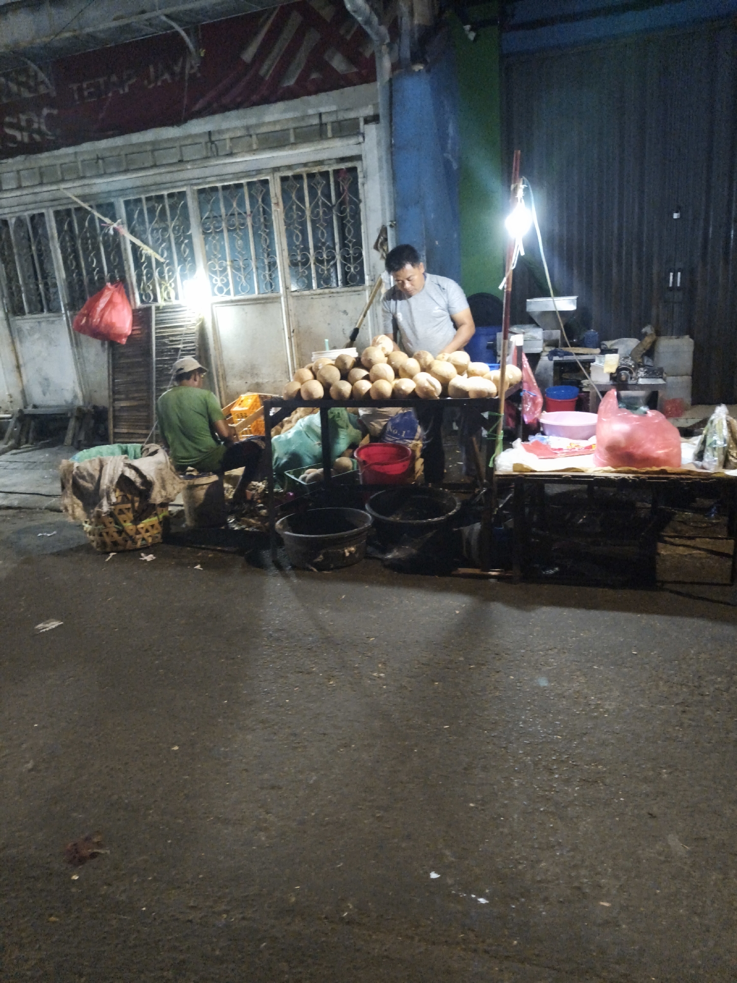 The image depicts a nighttime street scene in South Jakarta, Indonesia, featuring a street vendor selling coconuts. A man in a grey t-shirt stands behind a table laden with a large pile of coconuts, illuminated by a bright overhead light. To his left, another man wearing a green shirt and a cap sits on a bucket, facing away from the camera. Several large baskets and buckets are visible on the ground, some containing coconuts or other goods. In the background, a storefront with barred windows and a red banner with Indonesian text is present. The street is paved and appears wet, possibly from recent rain. The overall atmosphere suggests a typical bustling street market at night.