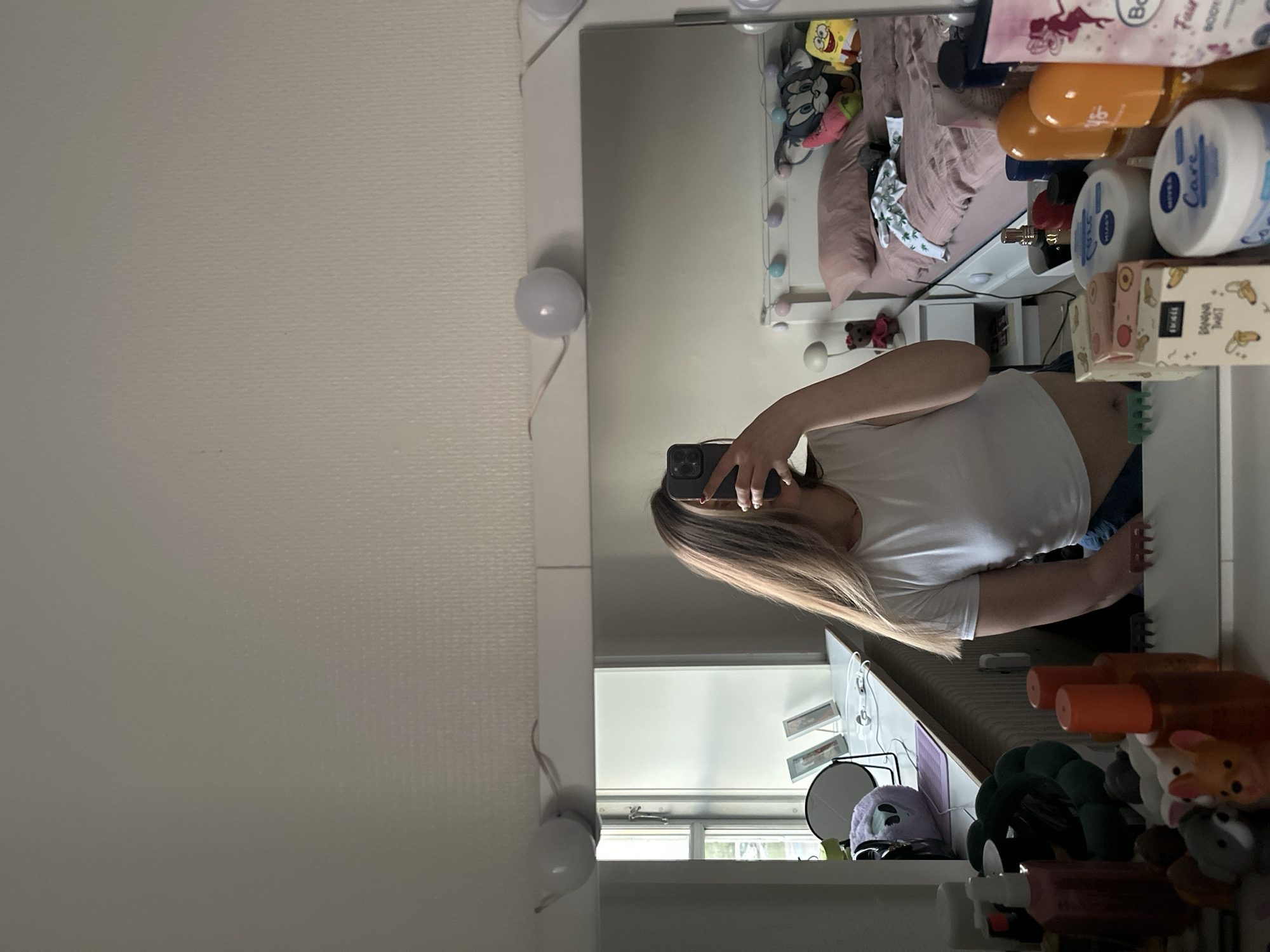 A woman is taking a picture of herself in a mirror. The mirror is in a bedroom. The woman is wearing a white t-shirt and jeans. The bedroom has a bed, desk, and window. The desk is cluttered with beauty products and stuffed animals. Based on the interior features and general context, it is inferred that the location is potentially a residence in Horsens, Denmark, although this cannot be definitively confirmed from the image alone.