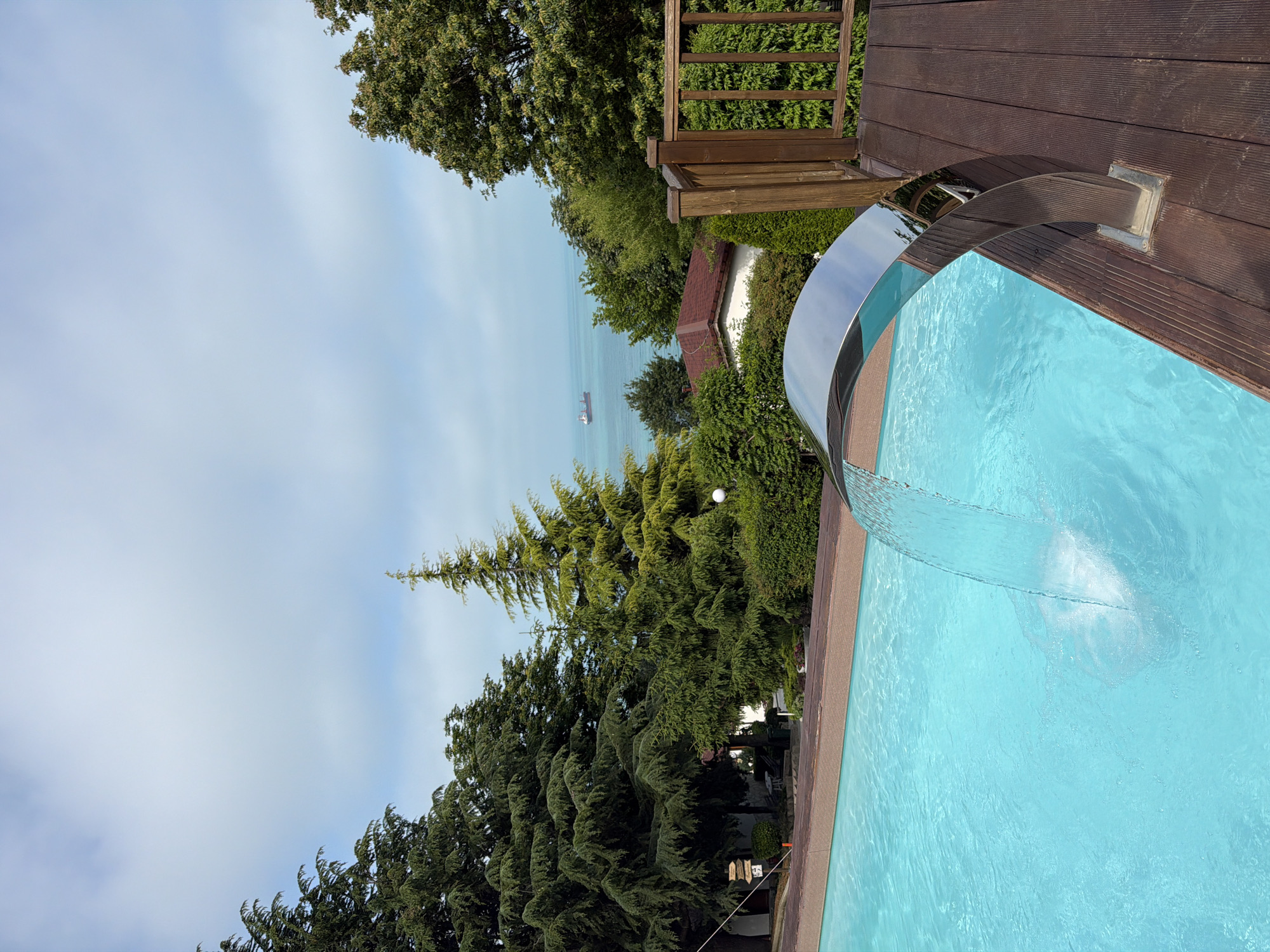 The image, when viewed in its correct horizontal orientation, captures a serene outdoor scene featuring a swimming pool with a distant view of the sea. In the foreground and mid-ground on the right, a bright blue swimming pool is visible, with a modern, curved chrome-like water feature gracefully pouring a continuous stream into the water, creating a gentle splash. Adjacent to the pool, a well-maintained dark wooden deck and a matching wooden railing frame the upper right portion of the scene.
Beyond the pool and deck, a rich natural landscape unfolds. Numerous dense green trees, including tall, conical evergreens and varied broadleaf trees, dominate the left side and background, providing a lush, private setting. Amongst the foliage, a small section of a building with a distinct red roof is partially visible. High above the distant tree line, the calm, light blue expanse of the Black Sea stretches towards the horizon, where a cargo ship or similar vessel appears as a small, faint silhouette. The sky above is a clear, pale blue with a few wispy clouds, suggesting bright, calm daytime weather, likely morning or early afternoon.
Notable details include a small white spherical object suspended against the green backdrop of the trees, and some decorative elements resembling wind chimes hanging near the base of the trees. The overall impression is one of tranquility and natural beauty, characteristic of a scenic coastal property in Varna, Bulgaria. There is no visible text in the image.