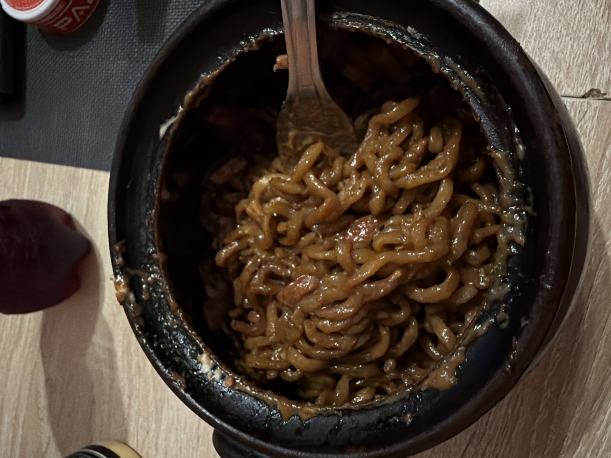 A close-up overhead shot shows a bowl of brown noodles being eaten with a fork. The noodles appear to be in a dark sauce. The bowl is black with a crusty edge. A can and a reddish-purple item are visible in the periphery. The table surface is a light wood grain. The location is unknown, and there's no discernible evidence to place it in Sofia, Bulgaria. There are no people present.