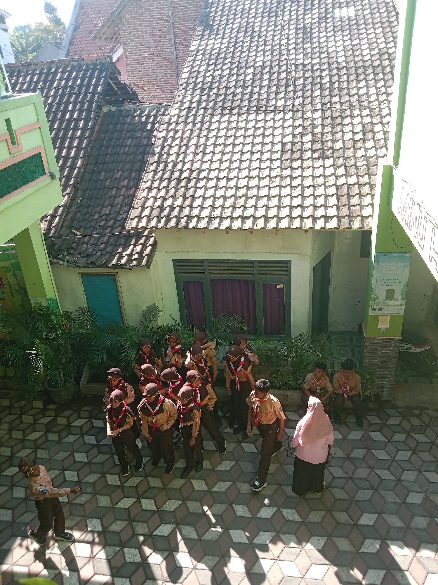 In Probolinggo, Indonesia, a group of approximately 15 Indonesian schoolchildren, all wearing brown scout uniforms with red and white neckerchiefs, are gathered outdoors on a patterned tiled courtyard. Most of the children are standing in a cluster, some facing towards the left of the frame, while others are looking at a female adult wearing a pink hijab and a dark skirt who is facing away from the camera. One child in the foreground, on the left side of the frame, is seen from behind and in profile, making a gesture with his right arm and looking towards the right, appearing to be interacting with the larger group. The environment includes the tiled courtyard, lush green potted plants and trees along a building with green window frames and dark purple curtains, and roofs of buildings in the background. Sunlight casts distinct shadows on the tiled ground, indicating a bright day. The buildings in the background are older structures with tiled roofs and brick elements. A banner with Indonesian text is visible on the right side of the building.