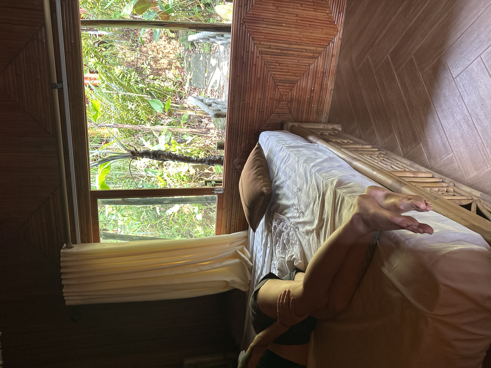 This image captures the interior of a room with a person relaxing on a bed, looking out onto a vibrant natural landscape. The room exudes a rustic and natural aesthetic, characterized by extensive use of wood and bamboo.
On the right, a person is lying on a bed with white sheets and a light brown pillow. Their lower body, specifically legs and midsection, are visible, suggesting a state of rest or relaxation. The bed features a decorative headboard or frame made of light-colored bamboo or wood, intricately arranged. The wall adjacent to the bed and extending towards the right is covered with light brown wood-paneled or tiled surfaces, arranged in a herringbone pattern. The ceiling, especially above the bed, is adorned with a unique bamboo paneling design, where individual bamboo sticks are arranged in a square, geometric pattern.
To the left, a large window offers a direct view of a lush, green outdoor environment, filled with dense foliage, ferns, and trees, indicative of a tropical or forested area. Bright natural light streams through the window, illuminating the vegetation outside, suggesting it is daytime with clear weather. Below the window, light-colored blinds or curtains are partially drawn, neatly folded upwards. The interior walls near the window are dark wood panels.
The overall scene conveys a sense of peacefulness and connection with nature. The materials used in the room's construction—bamboo, wood, and natural light—reinforce this serene atmosphere. There is no visible text in the image. The setting appears to be a natural retreat, consistent with the location context of Thông Nguyên Commune, Vietnam.