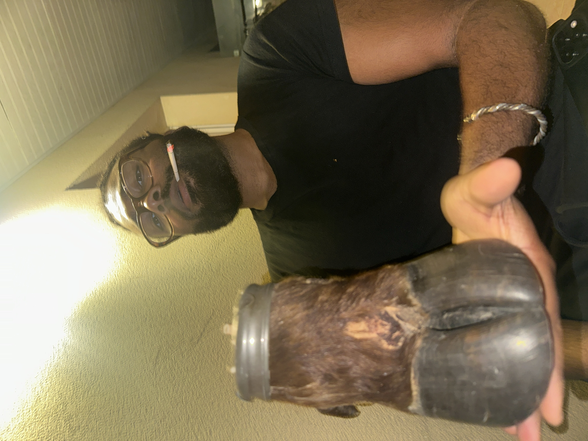 The image shows a dark-skinned man with a beard and glasses, holding what appears to be a taxidermied animal hoof. He is wearing a black t-shirt and a silver bracelet. A cigarette is visible behind his ear. The background is a light-colored wall, possibly with vertical grooves. The image was taken in Montreuil, France.